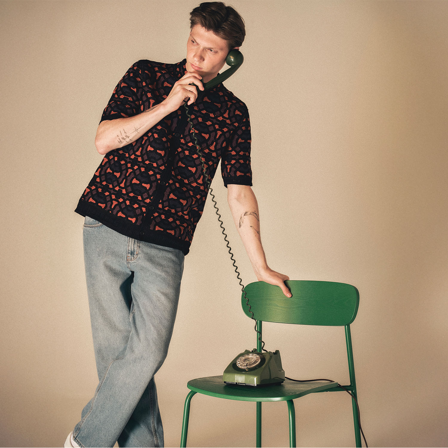 A person in a patterned shirt and jeans leans on a green chair while holding a vintage green rotary phone.