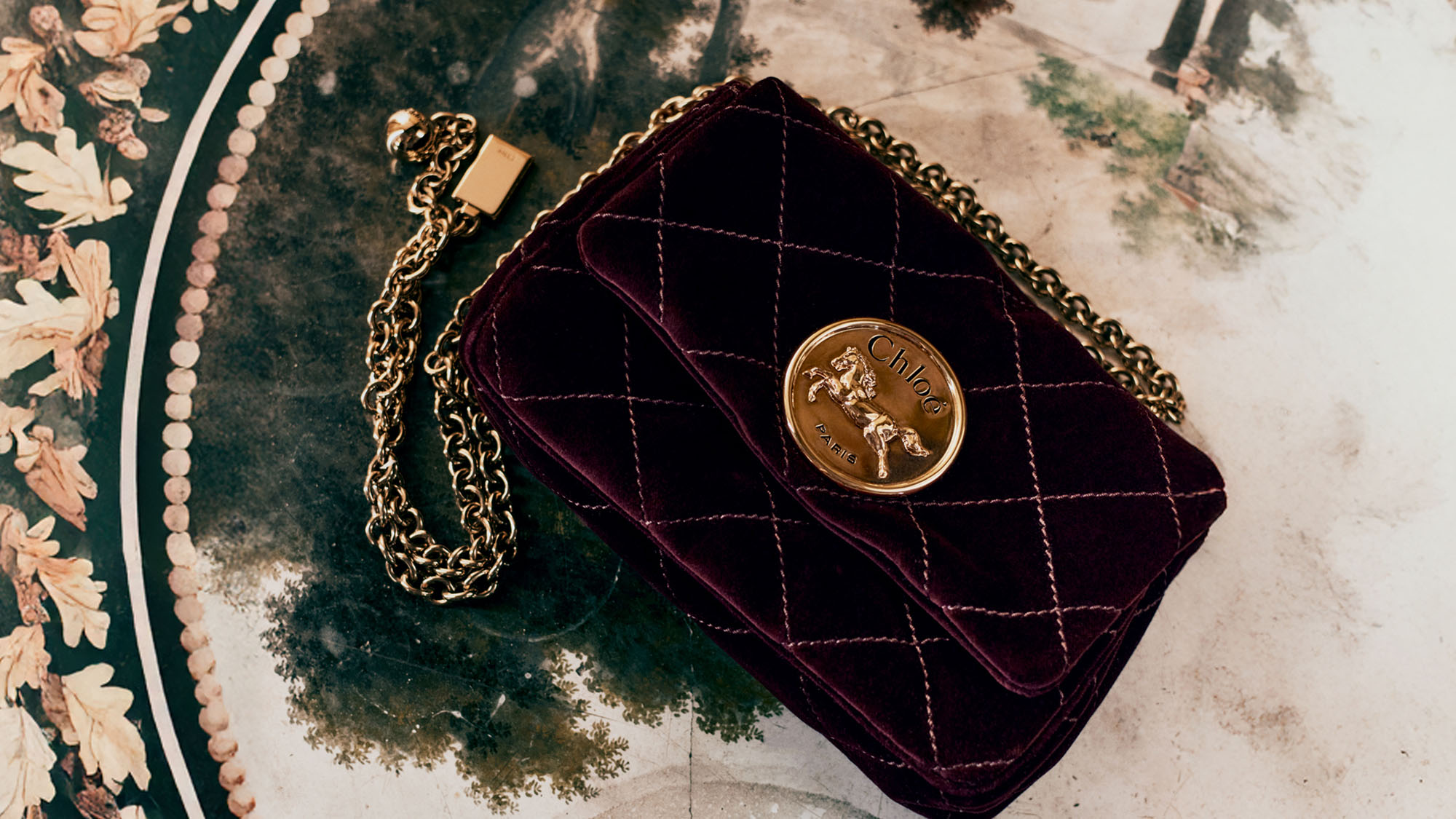 A quilted velvet purse with a gold chain and emblem lies on a decorative, painted table.