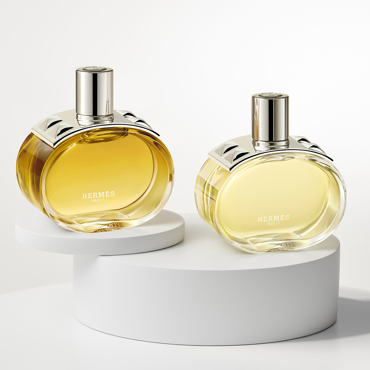 Two Hermès perfume bottles on white platforms, featuring elegant silver caps and yellow liquid inside.