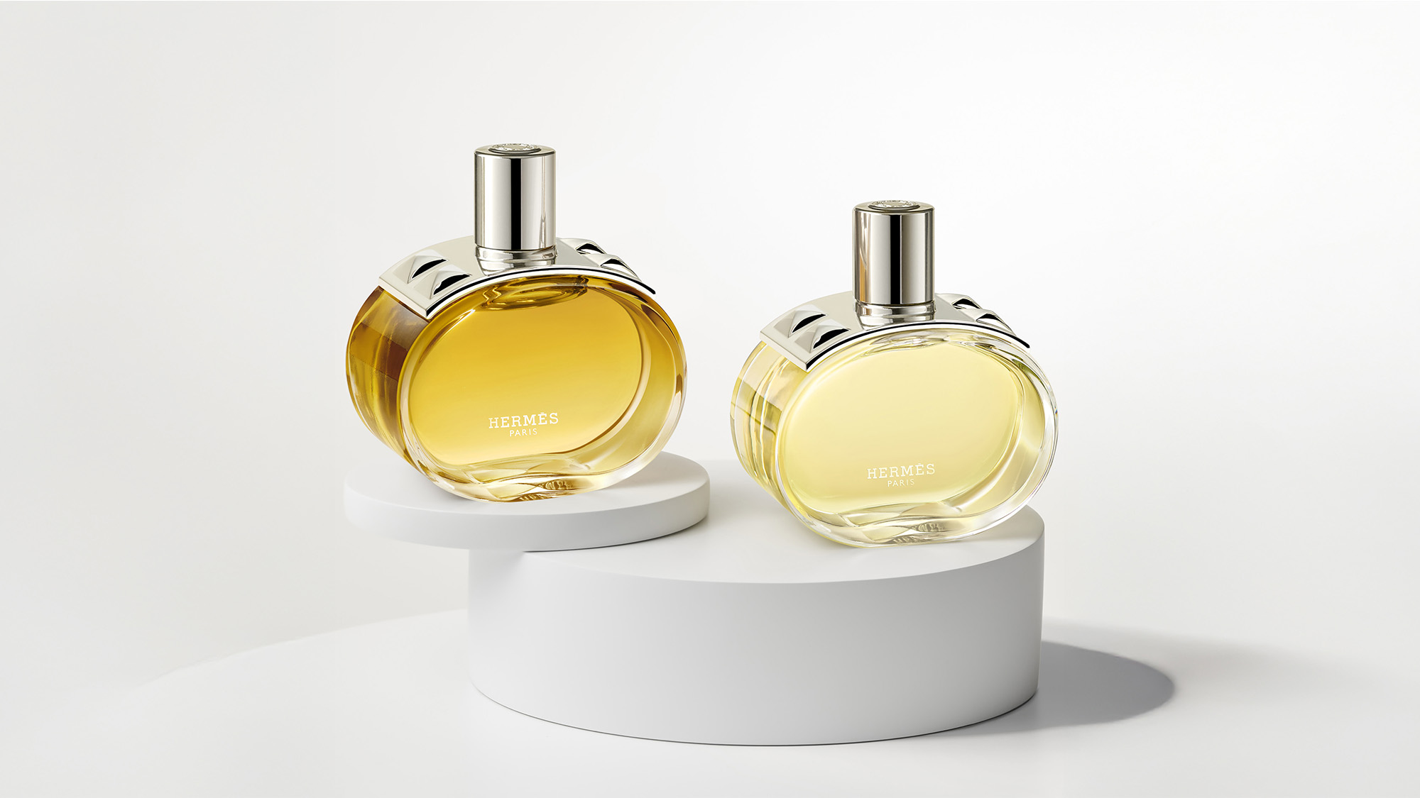 Two Hermès perfume bottles with silver caps displayed on white circular platforms.
