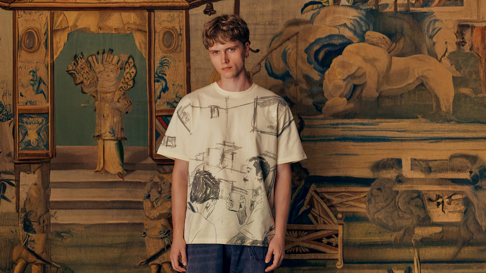A person stands in front of an ornate tapestry, wearing a graphic T-shirt and jeans.