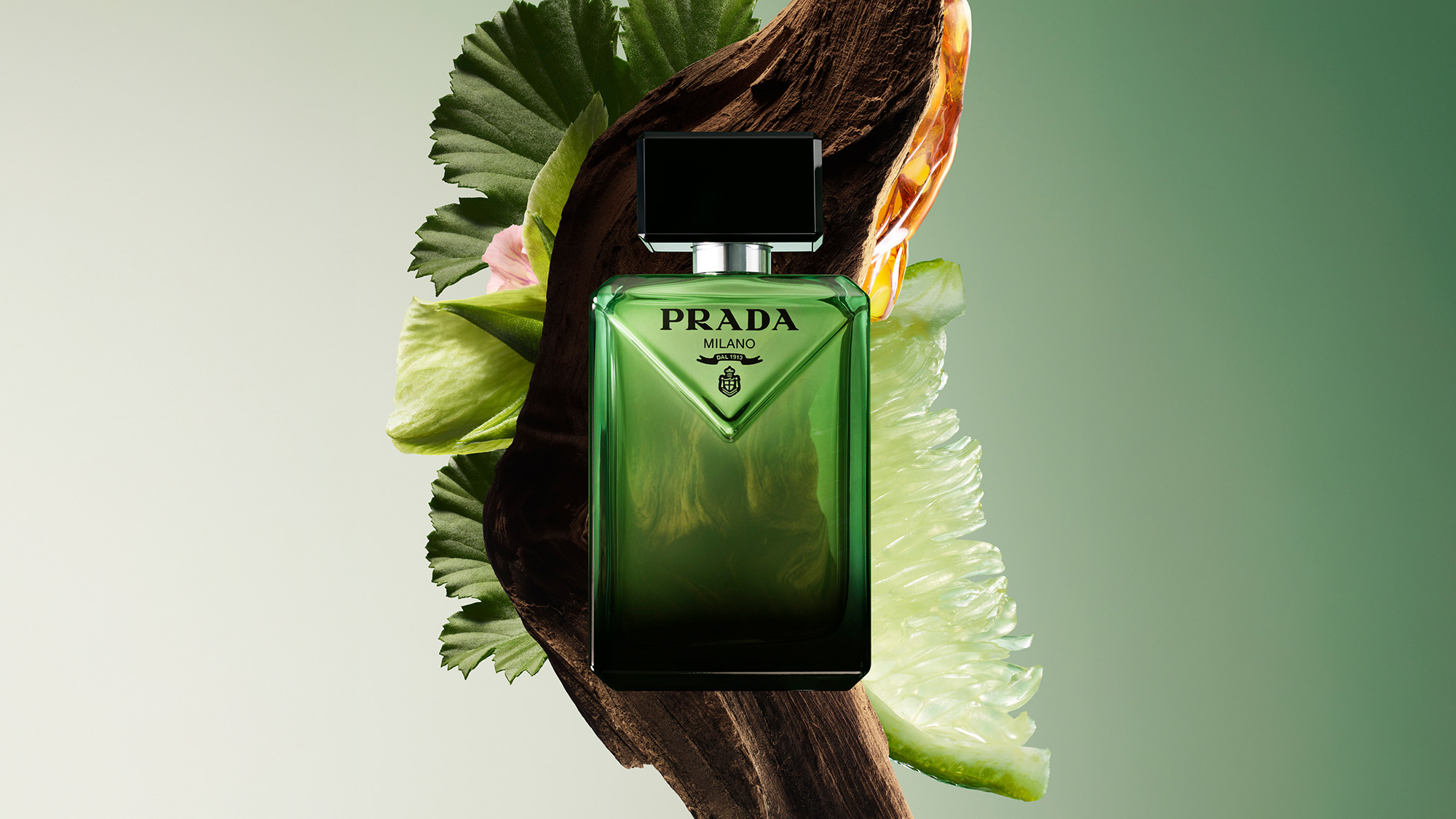 A green Prada perfume bottle with a black cap, surrounded by foliage and a large citrus slice against a light background.