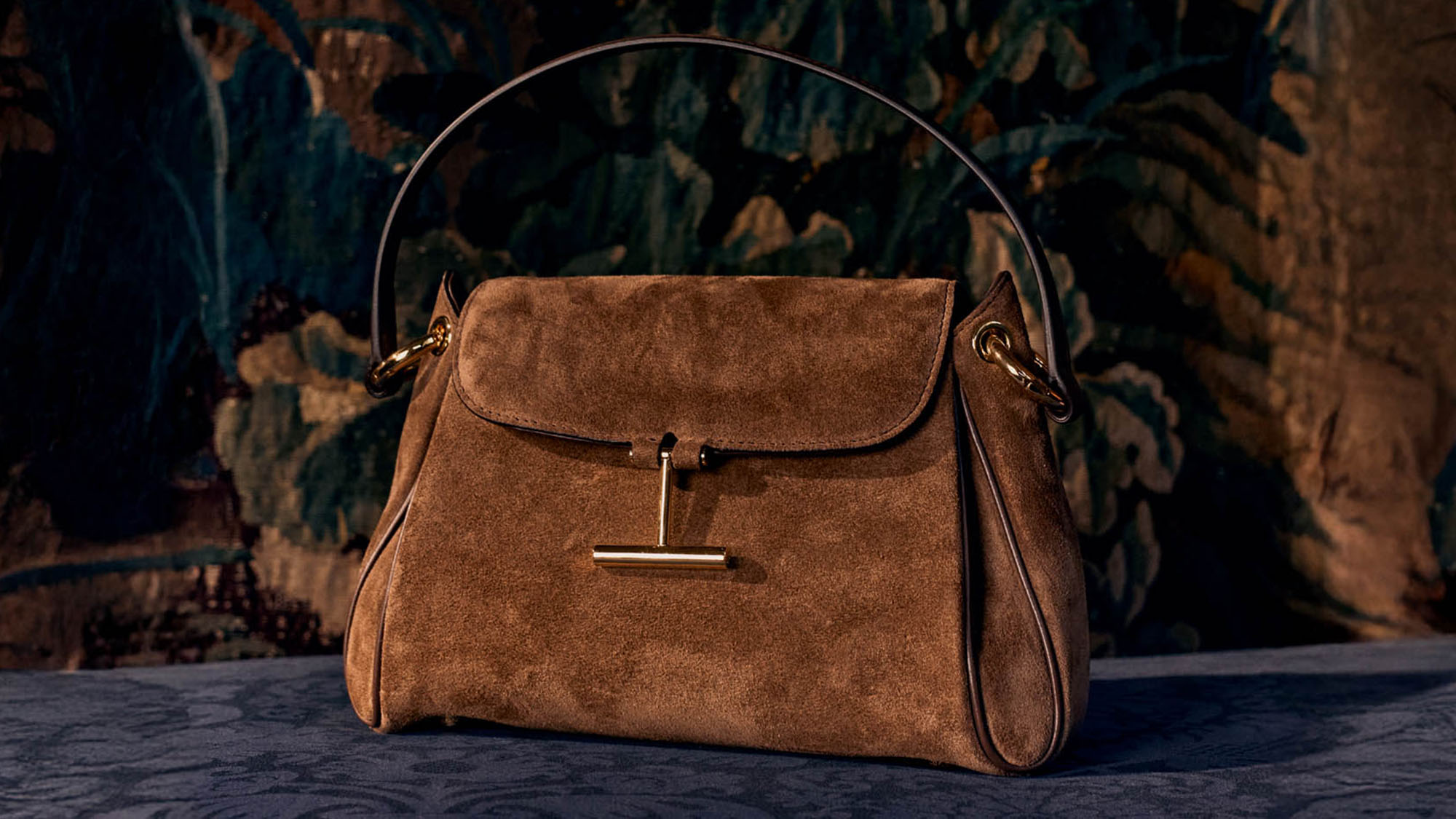 A brown suede handbag with a gold clasp and handle sits on a patterned blue surface against a dark floral background.