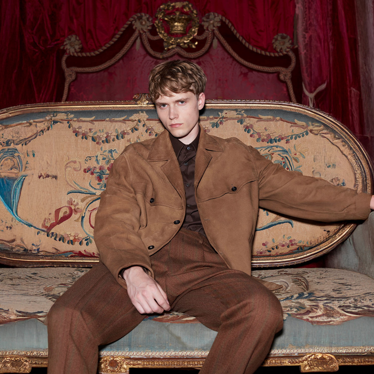 A person in a brown outfit sits on an ornate, floral-patterned sofa with a regal red and gold background.
