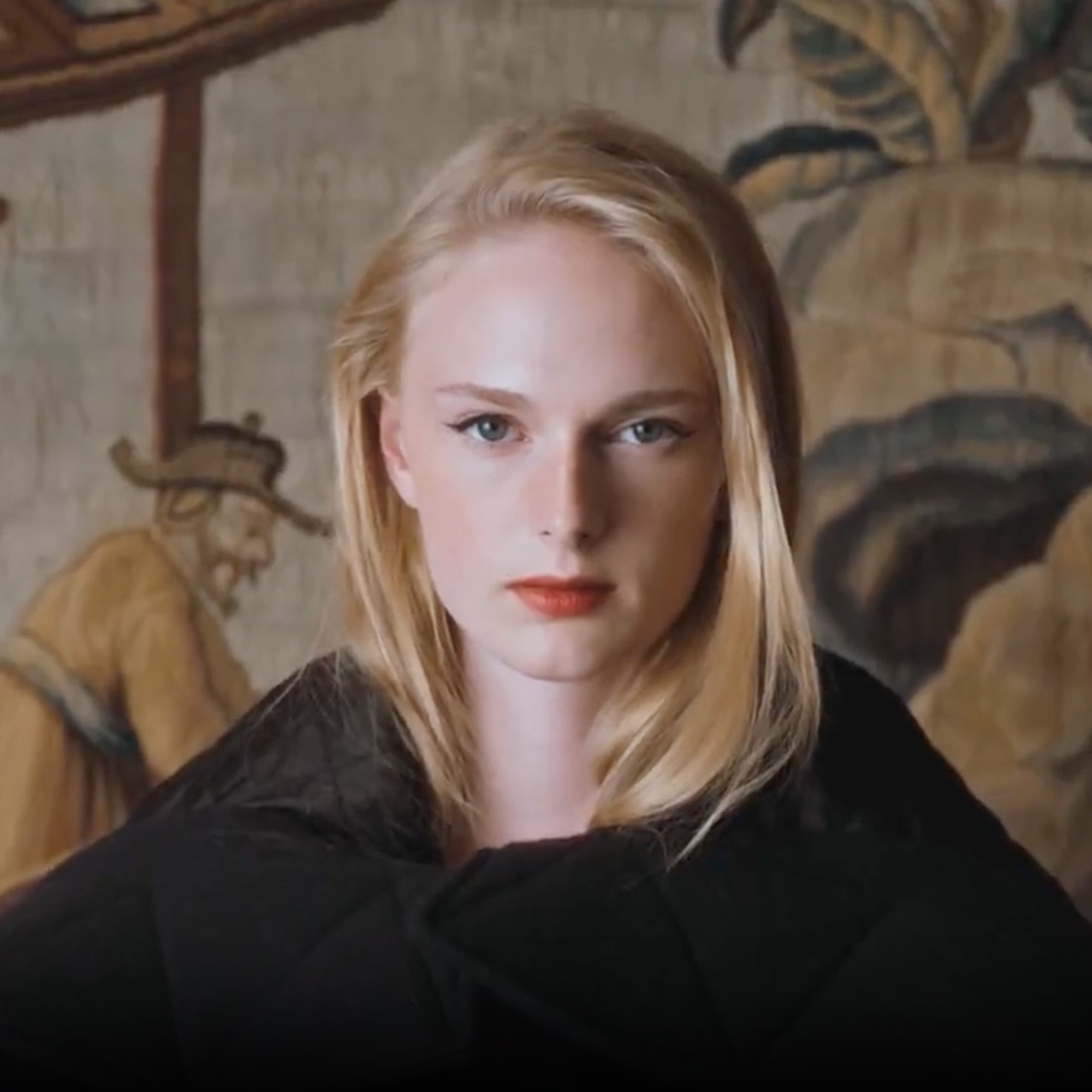 A person in a navy jacket and checkered skirt stands by a green chair with a tapestry backdrop.'
