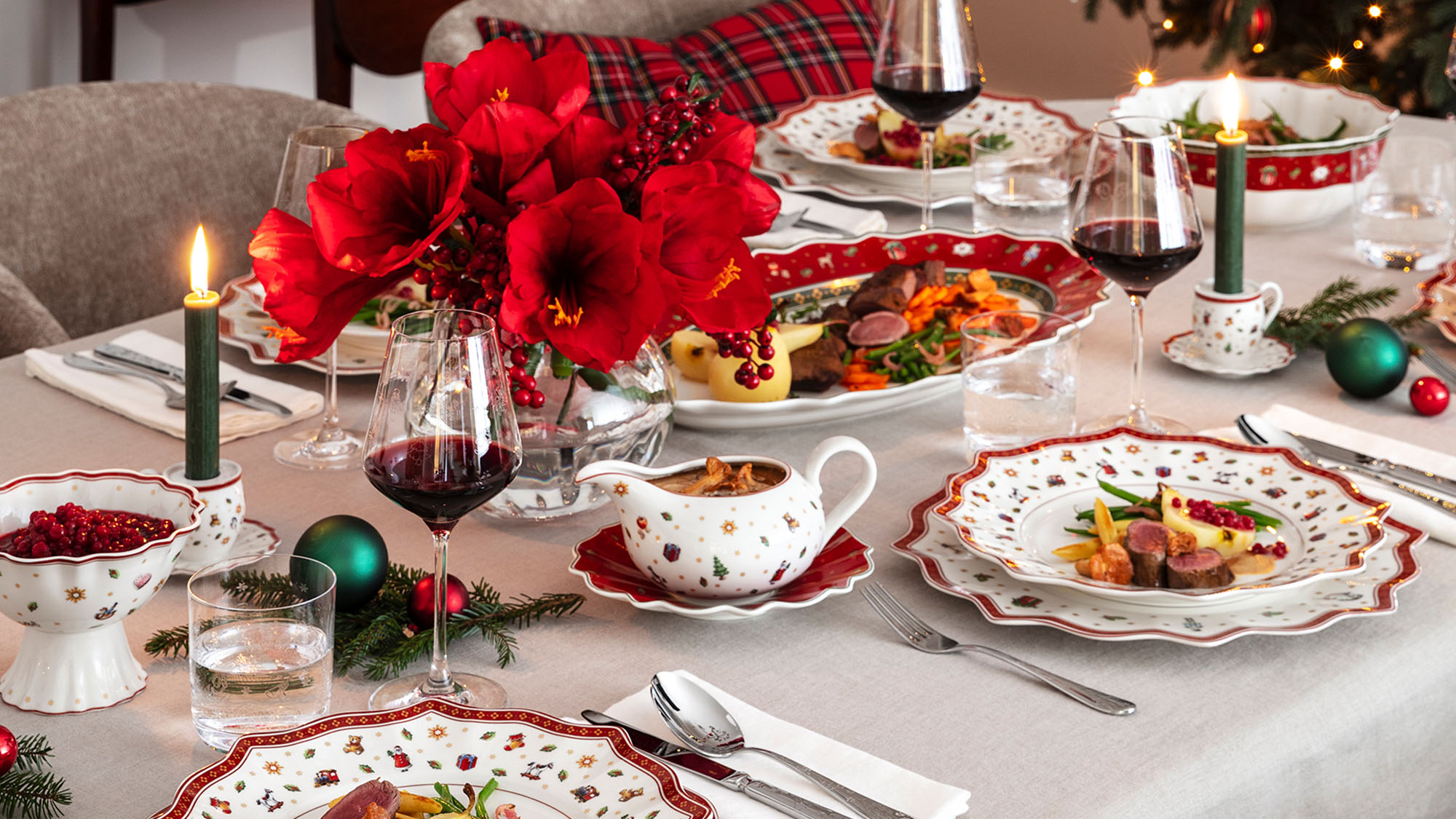 Elegant holiday dining table with red and white plates, red flowers, lit candles, and wine glasses. A Christmas tree glows softly in the background.