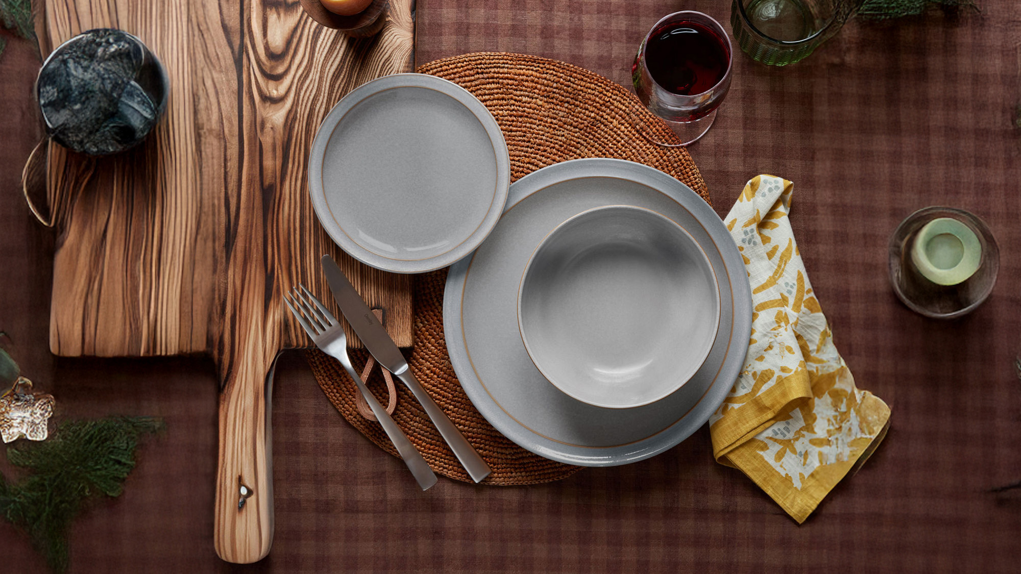 Table setting with gray plates on a woven mat, cutlery, candle, wine glass, and yellow-patterned napkin.