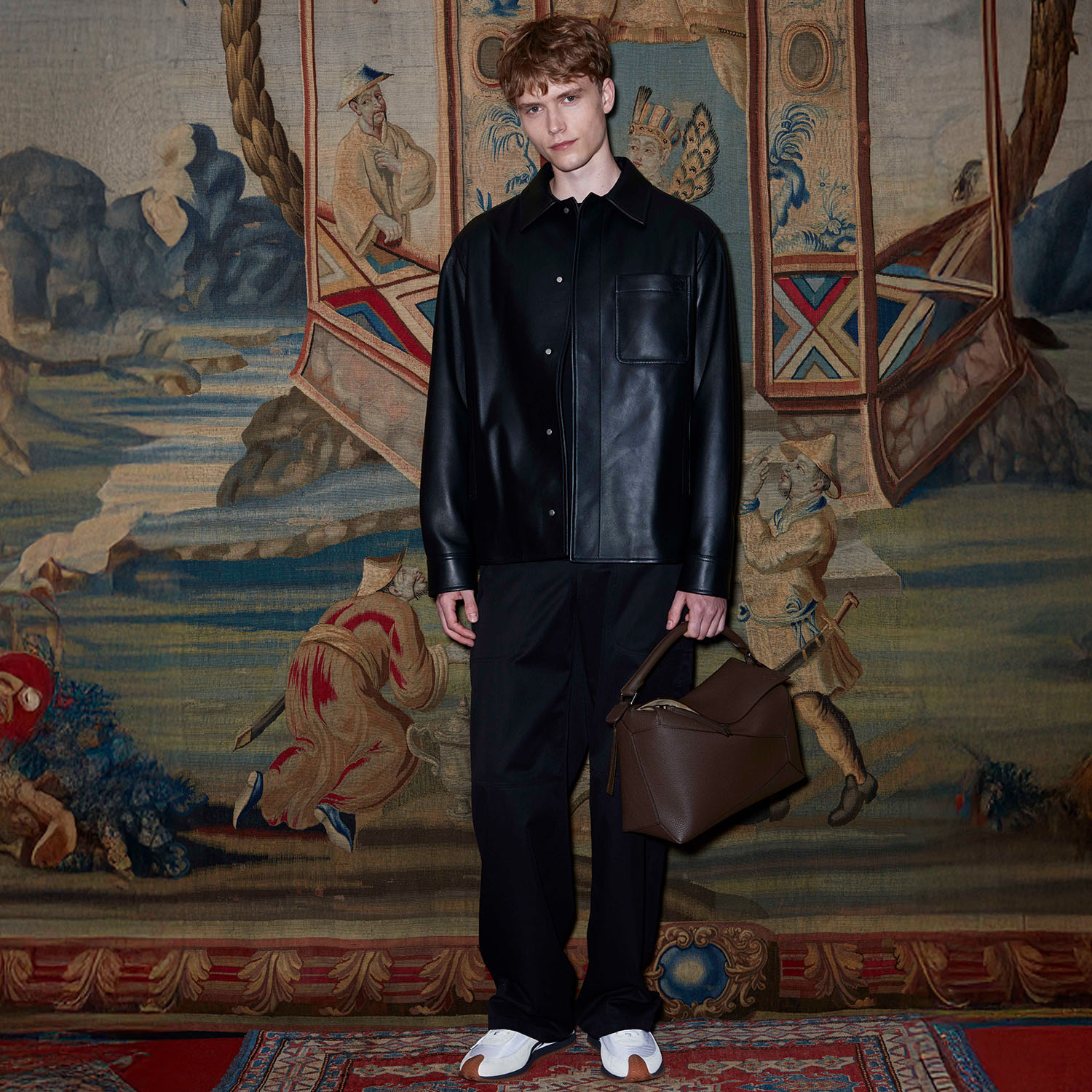 A person in a black leather outfit holds a brown bag, standing in front of a detailed tapestry with figures and elaborate patterns.