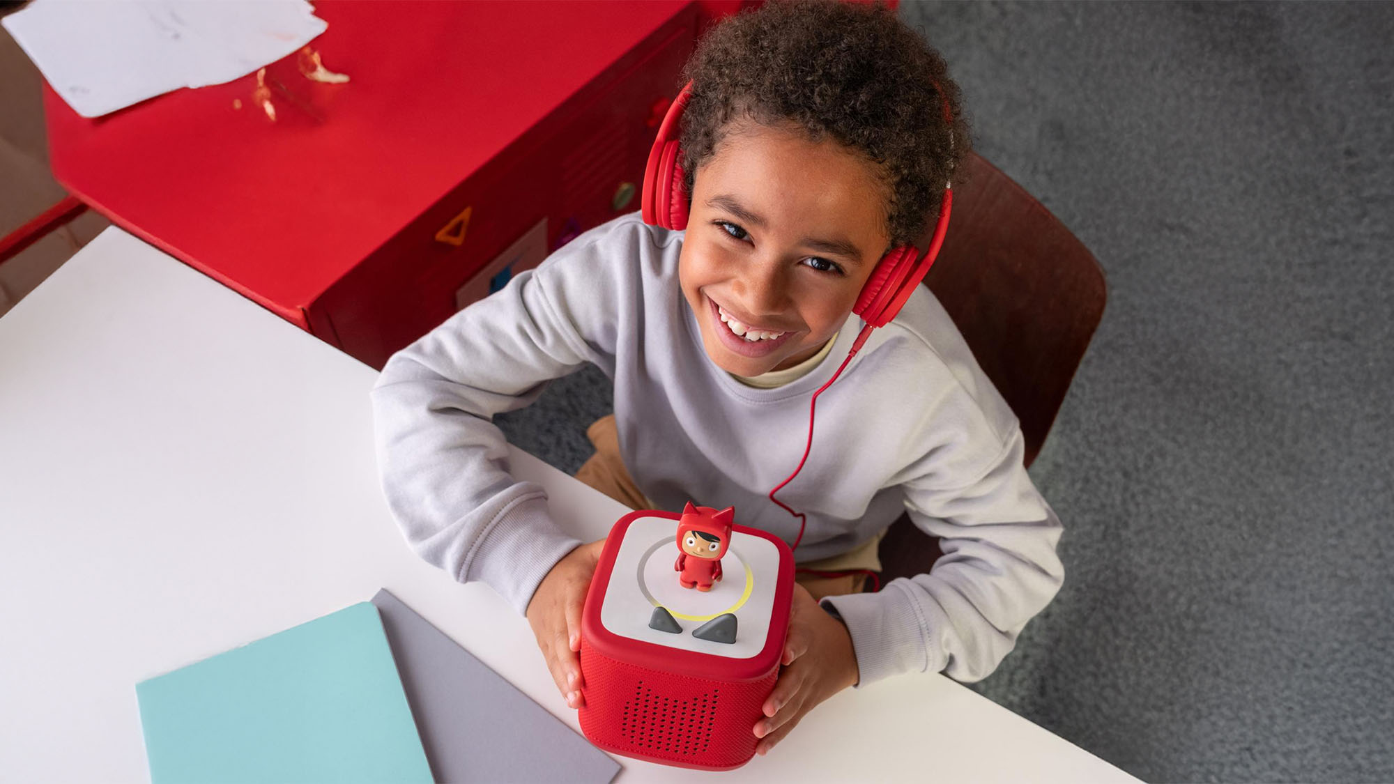 A child wearing red headphones smiles while using a red audio device with a small figurine on top.