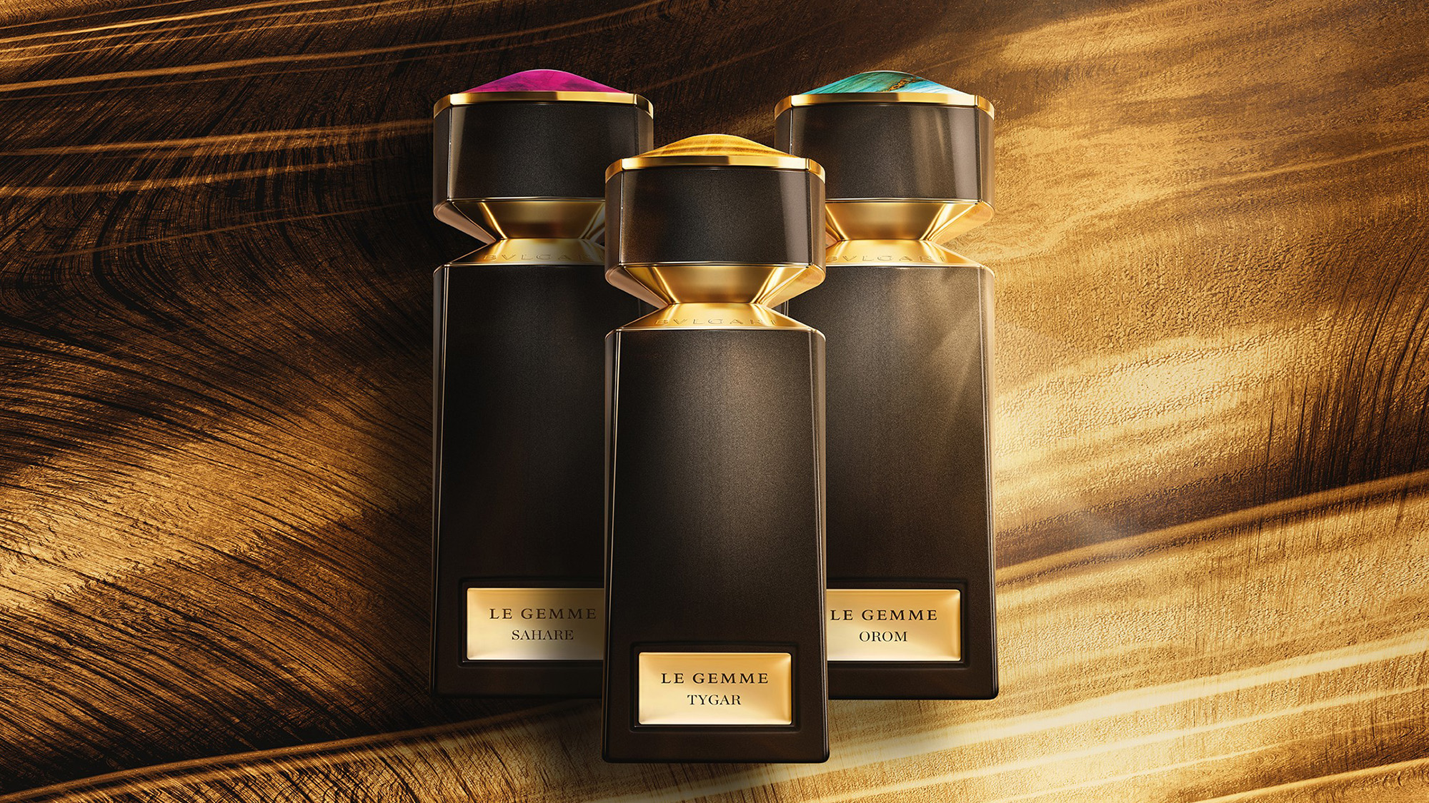 Three elegant perfume bottles with gold accents are displayed on a wooden surface, each topped with a distinct colored gem.