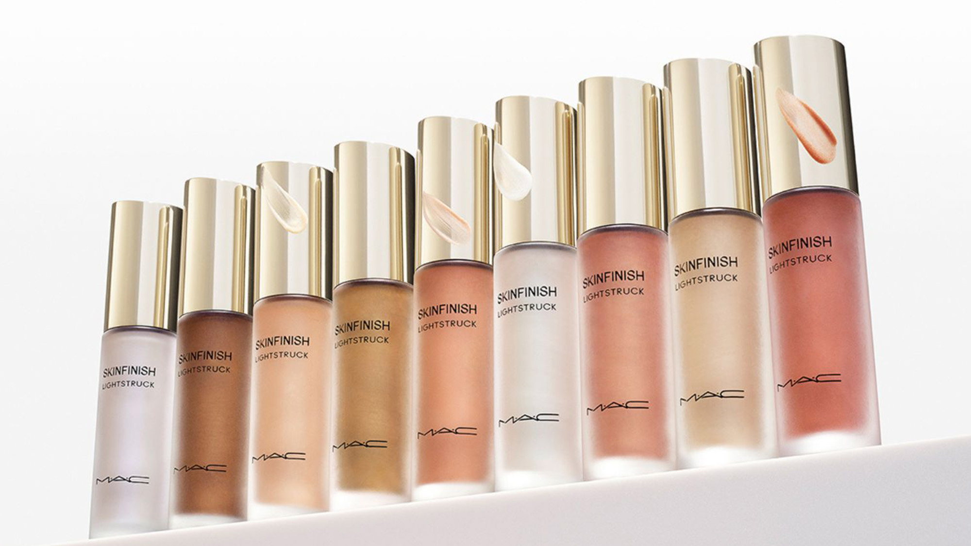 A row of MAC liquid highlighters in gradient shades with gold caps, displayed on a white surface.