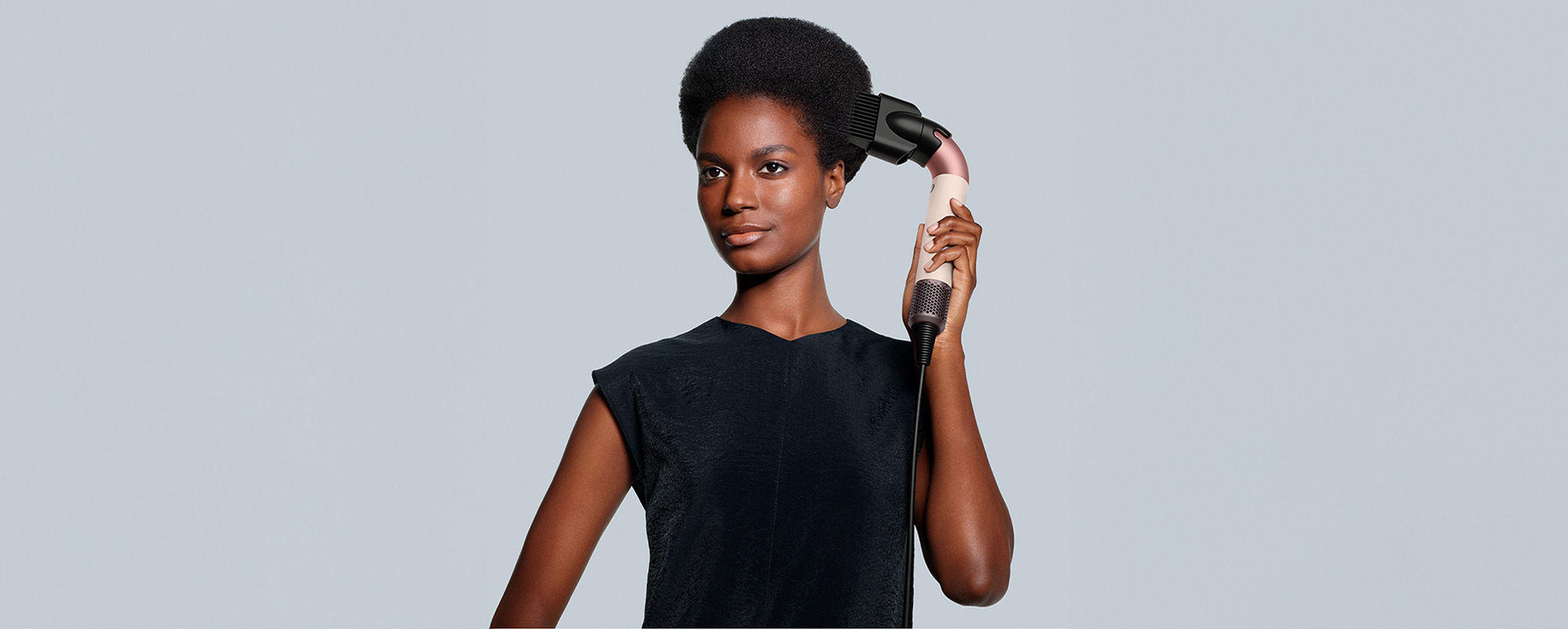 A person uses a hair styling tool on their hair while wearing a black top, set against a plain background.