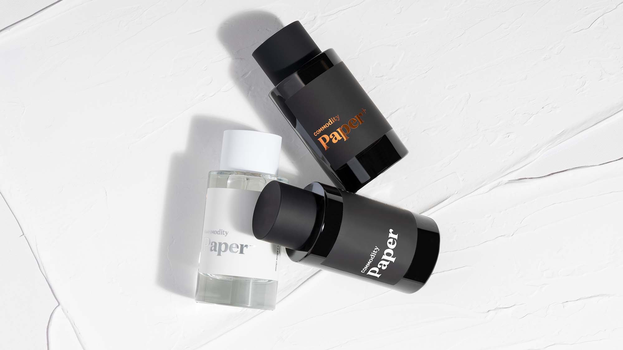 Three perfume bottles labeled 'Paper,' in black and white, arranged on white textured surfaces.