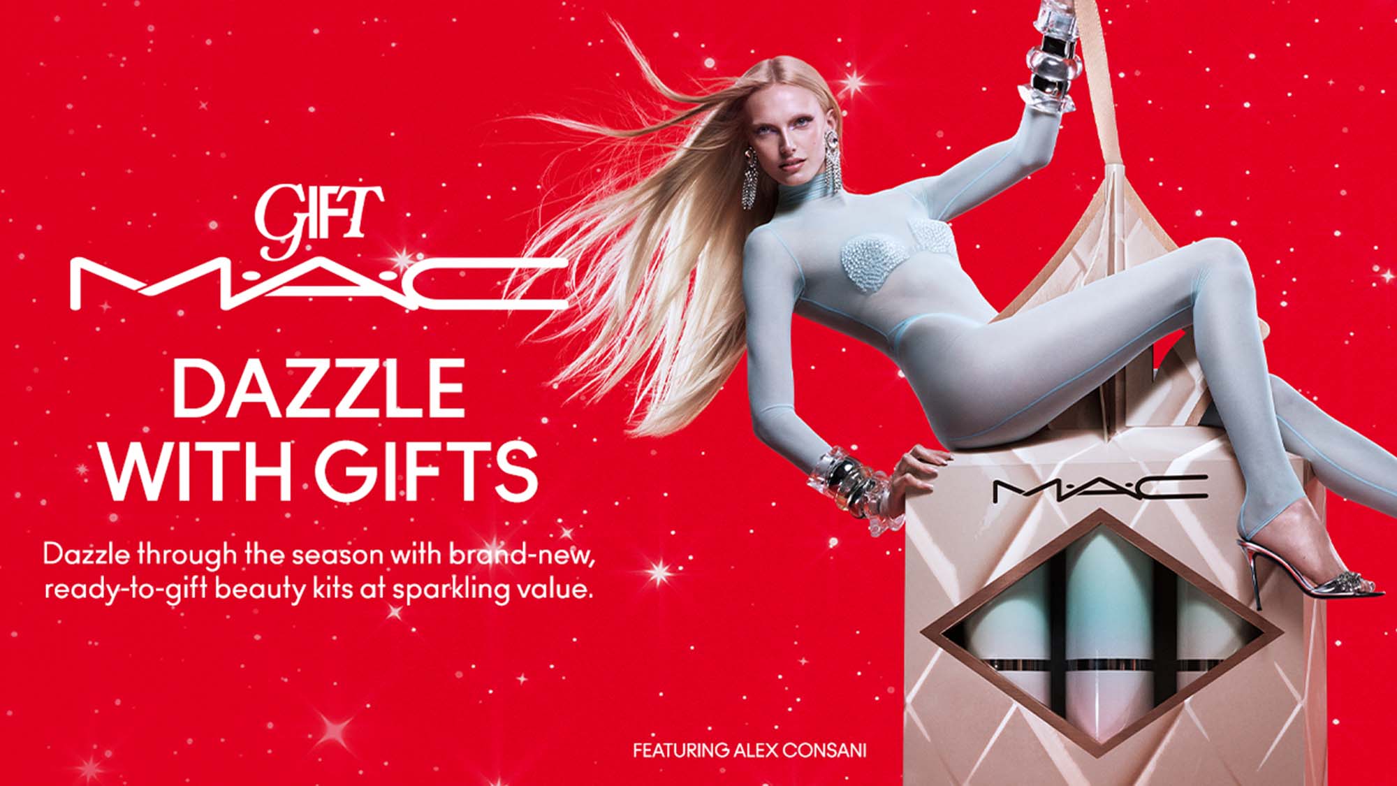 A person in a stylish outfit poses on a large MAC gift box against a red background, promoting beauty kits with the slogan 'Dazzle with Gifts.'
