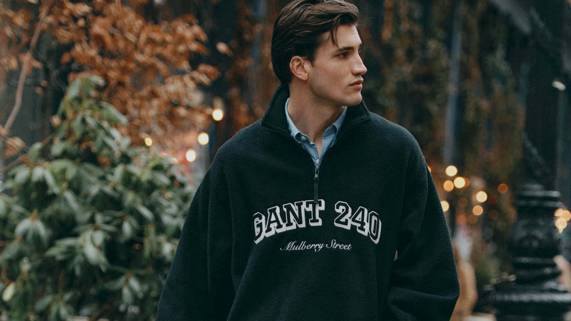A person in a dark sweater with 'GANT 240 Mulberry Street' looks to the side, outdoors, with autumn foliage and lights in the background.