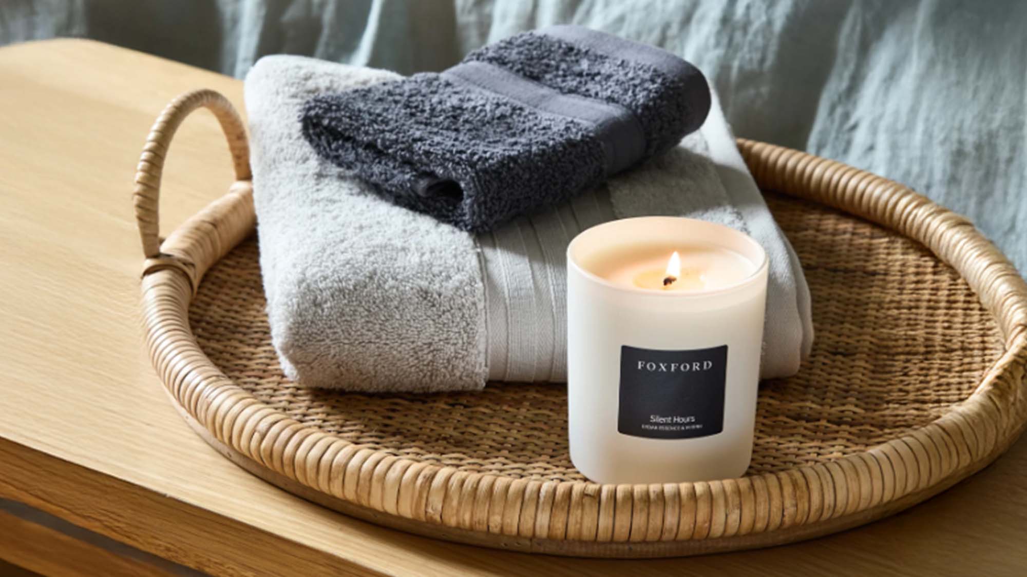 A Foxford candle, towels, and a rattan tray are arranged on a wooden bench beside a bed with blue bedding.