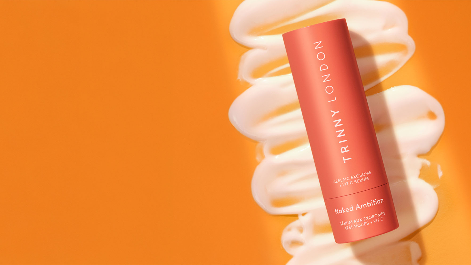 A coral-colored Trinny London bottle on an orange background with white cream streaks behind it.