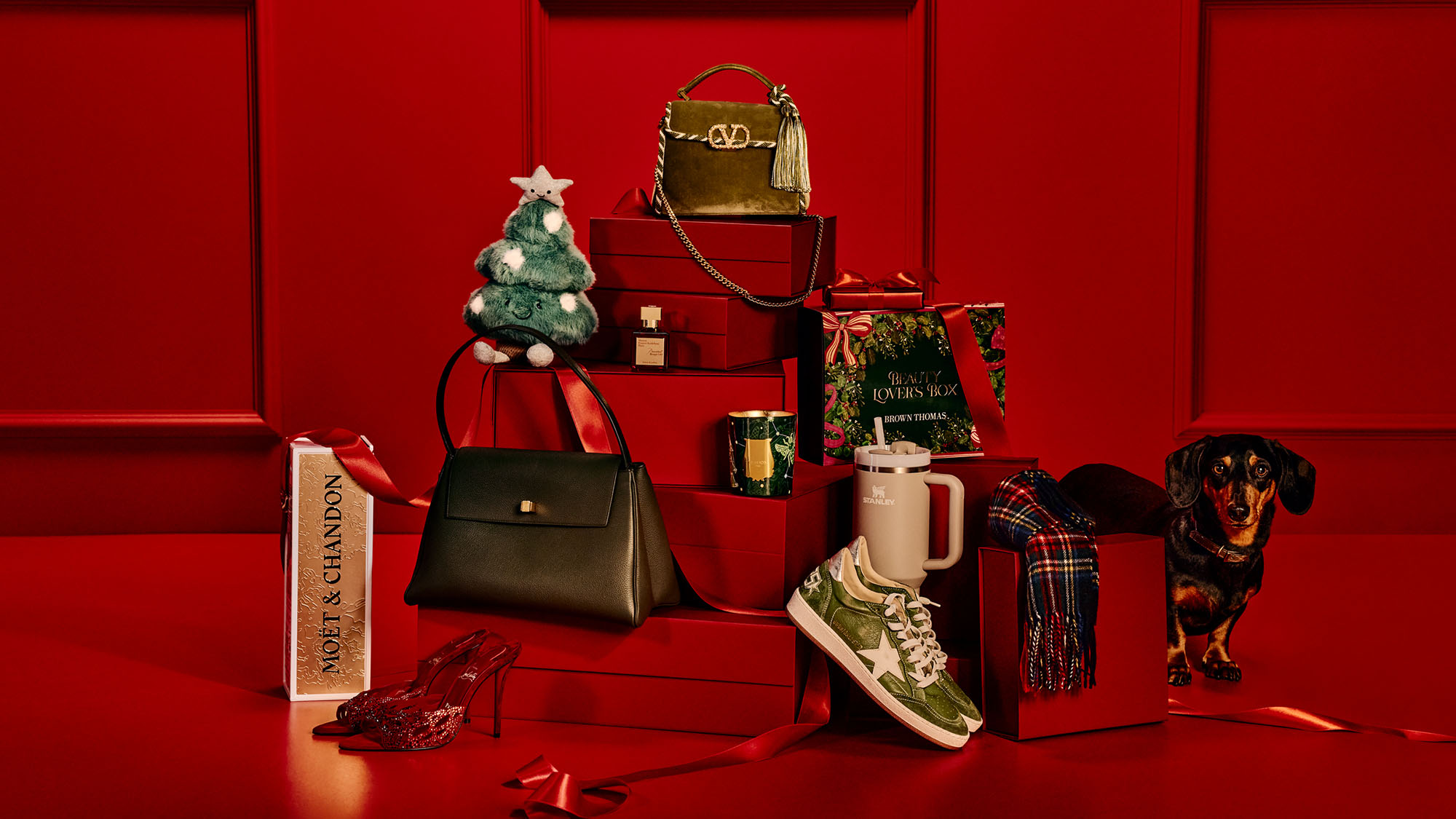A festive arrangement on a red background featuring luxury items: handbags, shoes, a small Christmas tree, and a dachshund figurine.