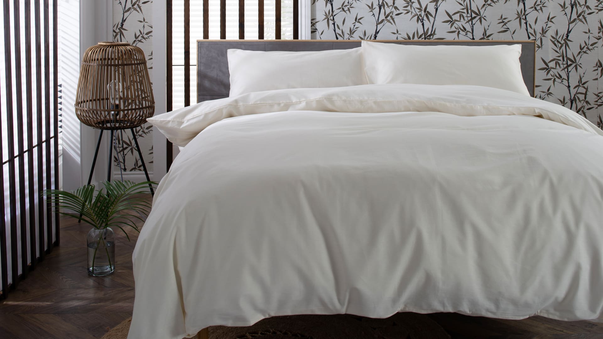 A neatly made bed with white linens in a room featuring a nature-themed wallpaper, a wicker lamp, and wooden flooring.