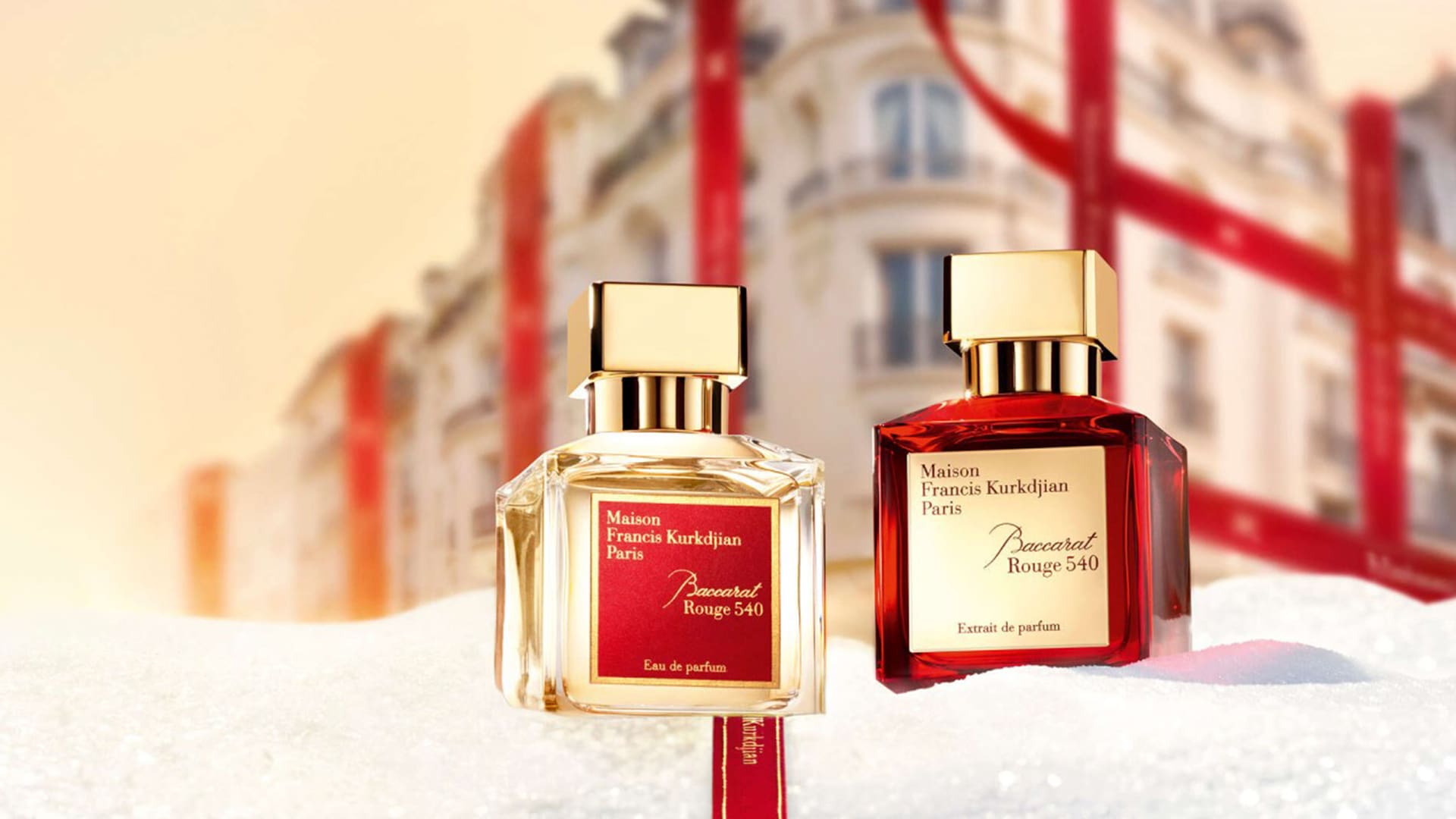 Two perfume bottles with gold caps sit on a snowy surface, with a festive, ribbon-decorated building in the background.