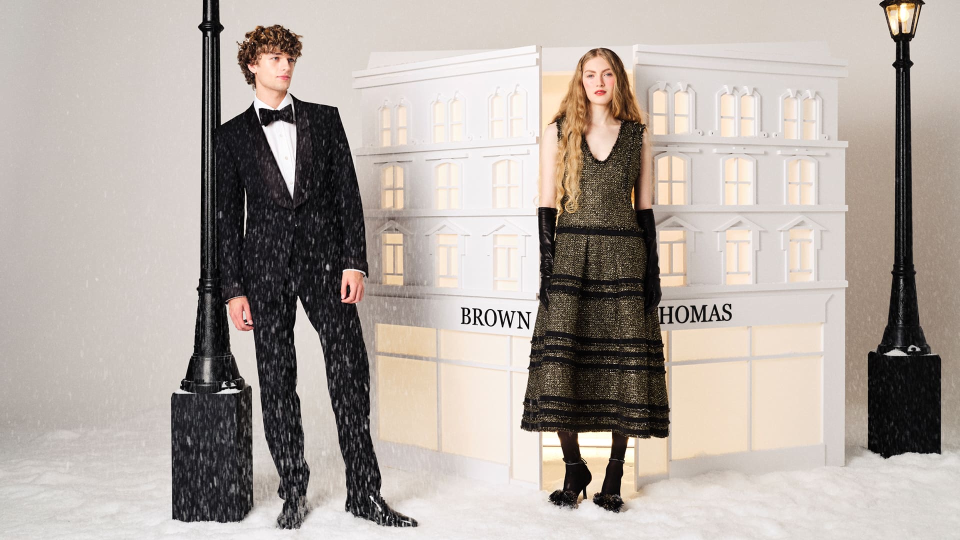 A man and woman in formal attire stand in a snowy scene with street lamps and a building facade labeled 'Brown Thomas.'