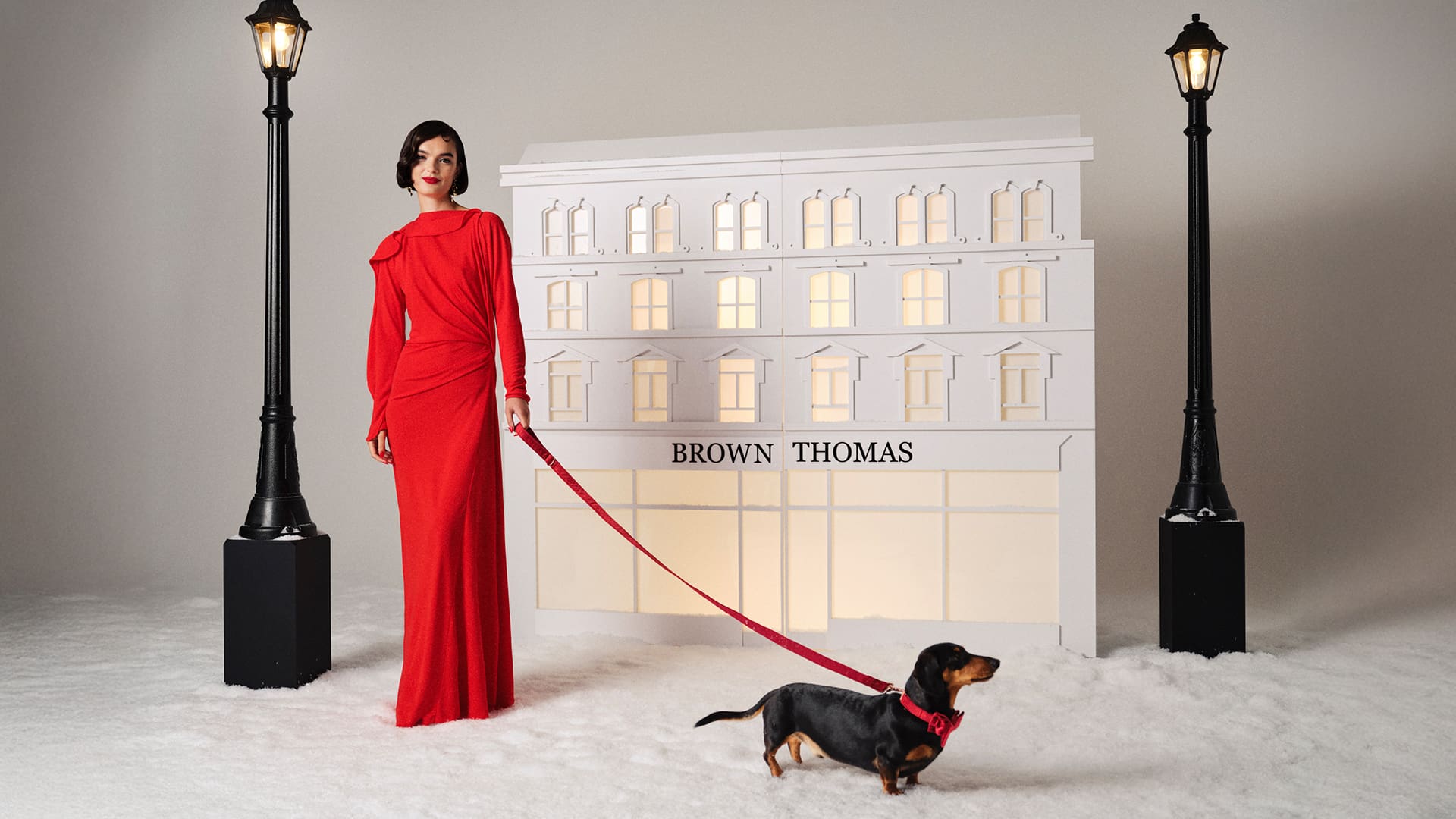A woman in a red dress stands with a dachshund on a red leash in front of a Brown Thomas storefront backdrop, flanked by two street lamps.