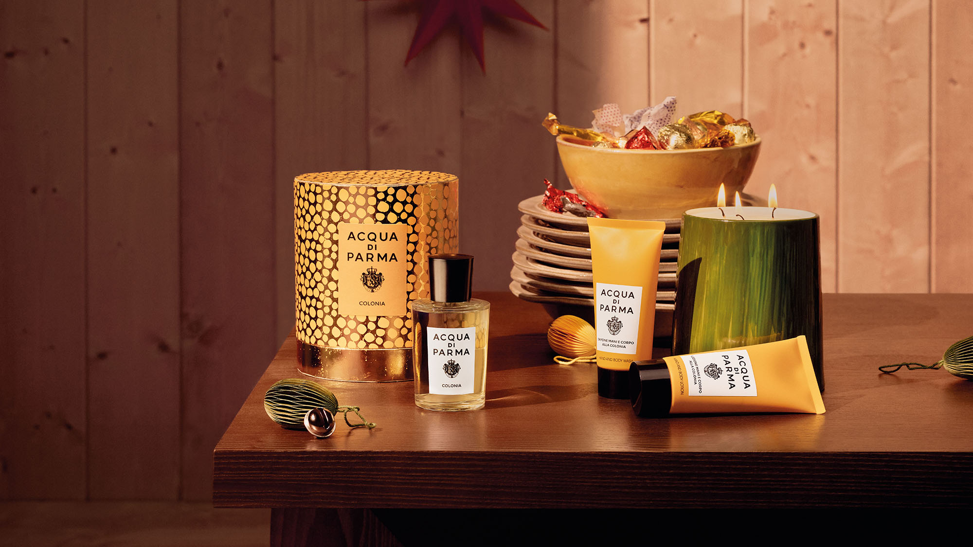 A festive table setup featuring Acqua di Parma products, a bowl of candies, and decorative ornaments on a wooden surface.