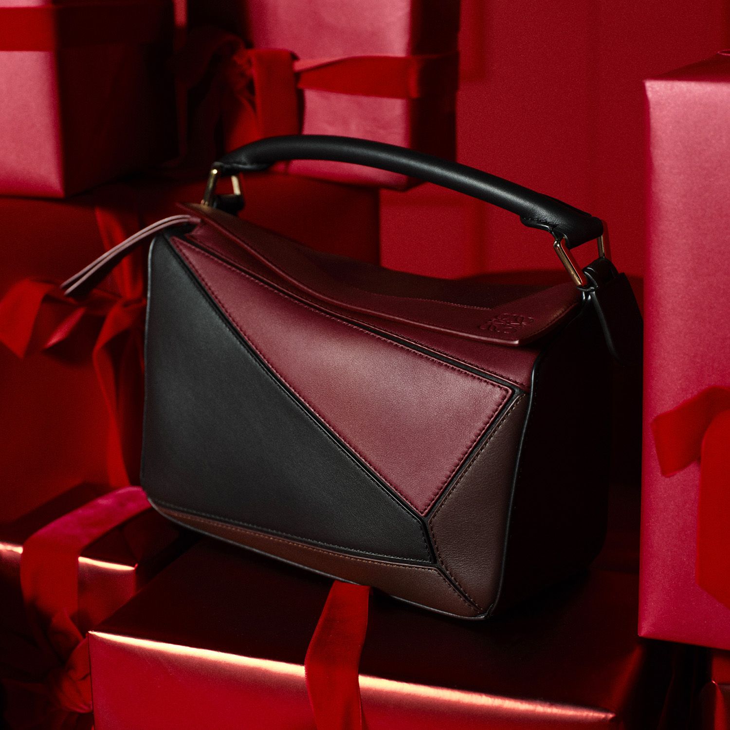 A Loewe handbag with geometric panels and a handle is set against red gift boxes tied with ribbons.