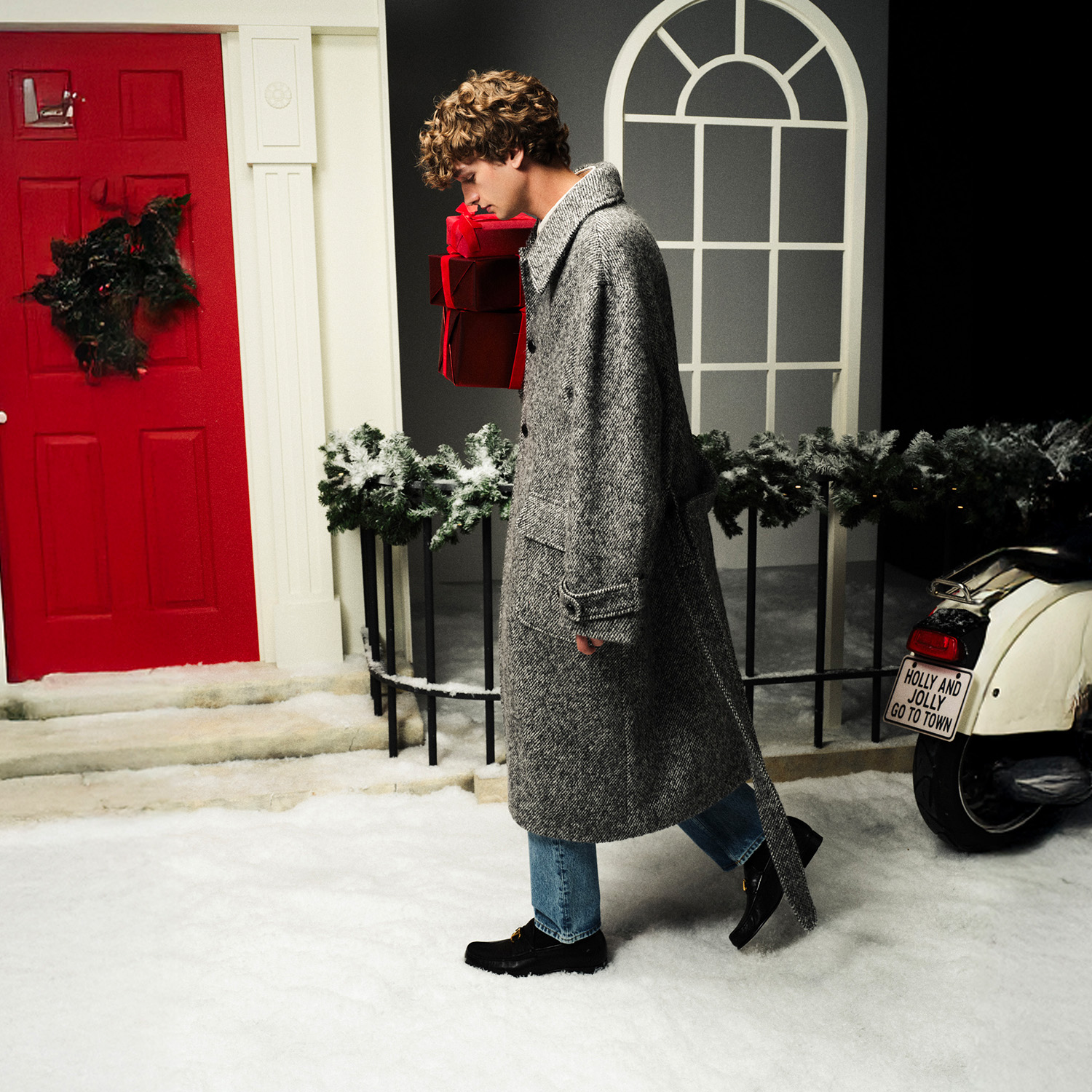 Person in a gray coat holds red gift boxes, walking on snow by a festive door and decor.