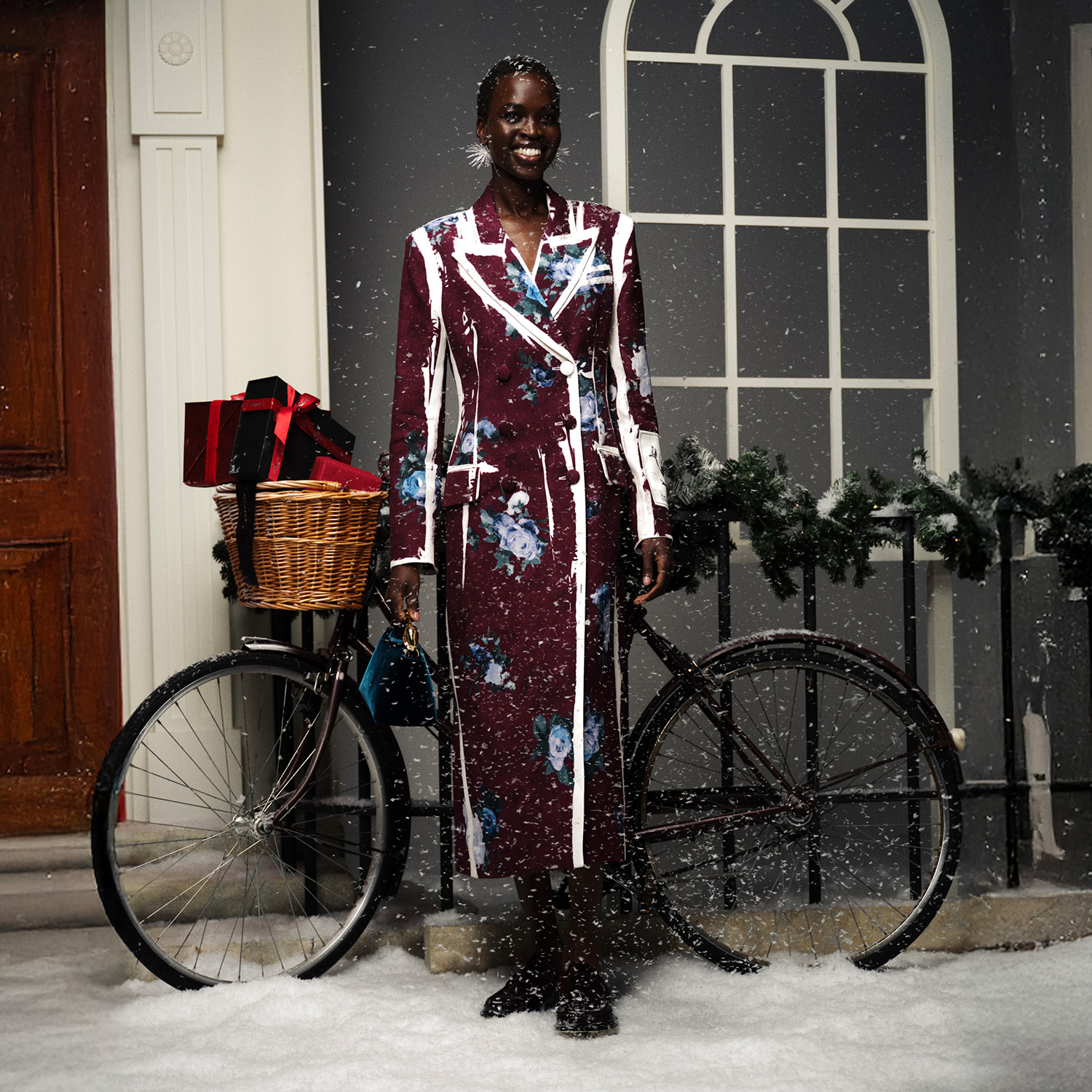 A person in a floral Erdem coat stands next to a bike with gifts in its basket, surrounded by snow and holiday decorations.