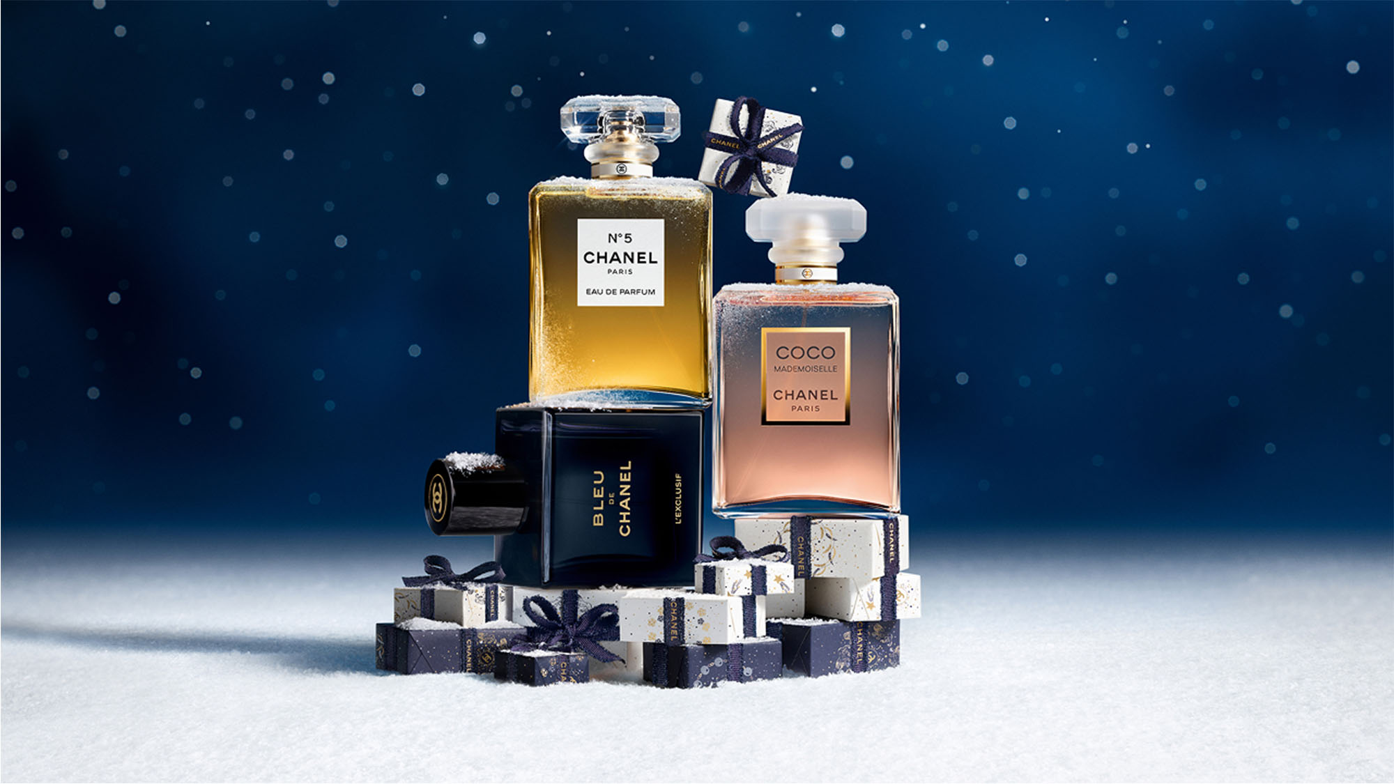 Perfume bottles of Chanel No. 5, Coco, and Bleu on a snowy surface with gift-wrapped boxes, set against a starry night background.