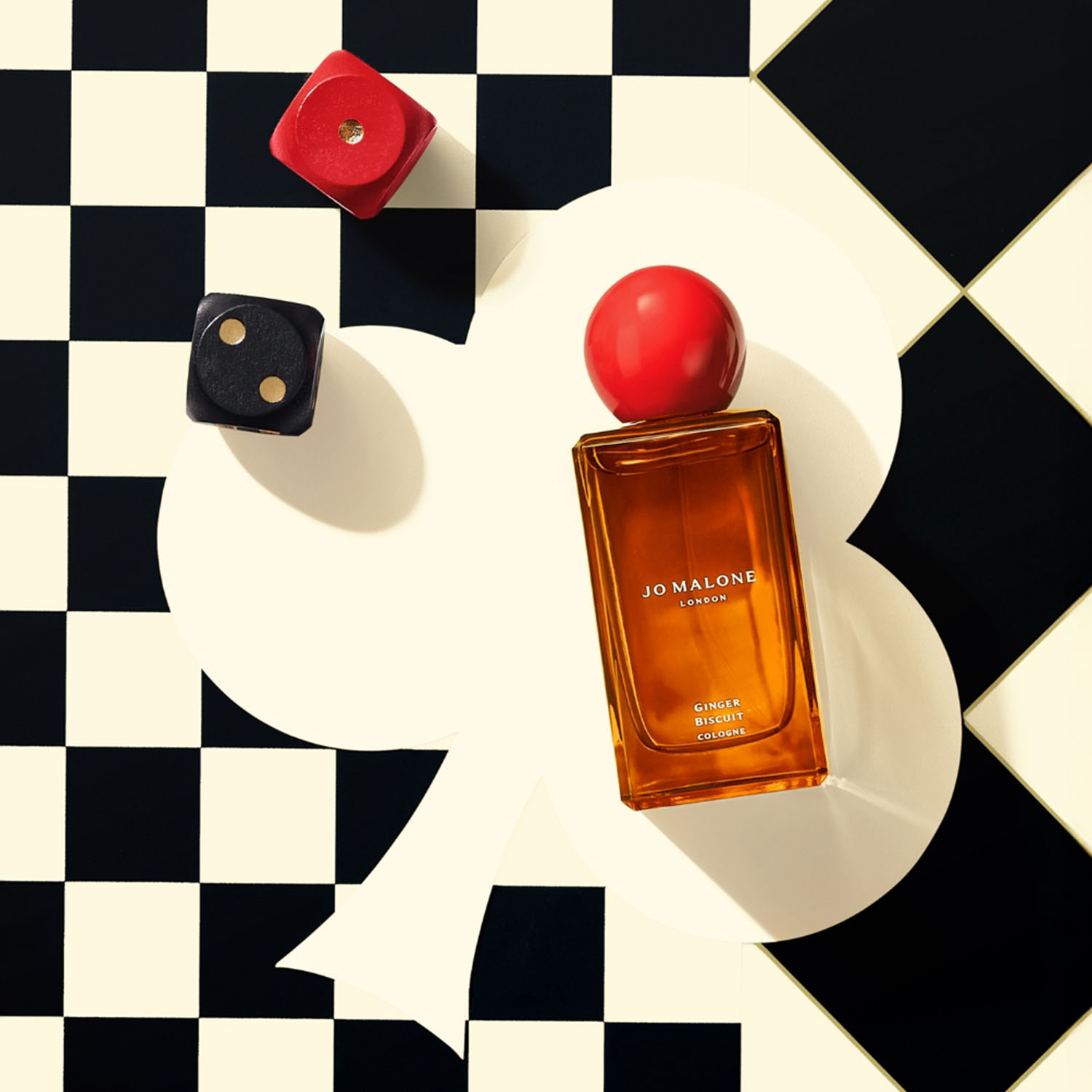 A Jo Malone perfume bottle with a red cap is placed on a black and white checkered background, accompanied by two dice.