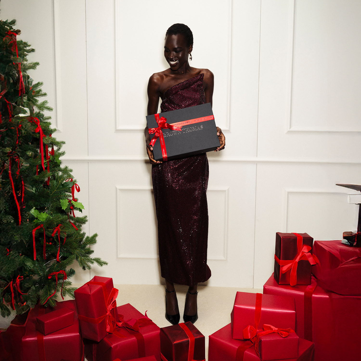 A smiling person in a sleek dress holds a gift box near a decorated Christmas tree and wrapped presents.
