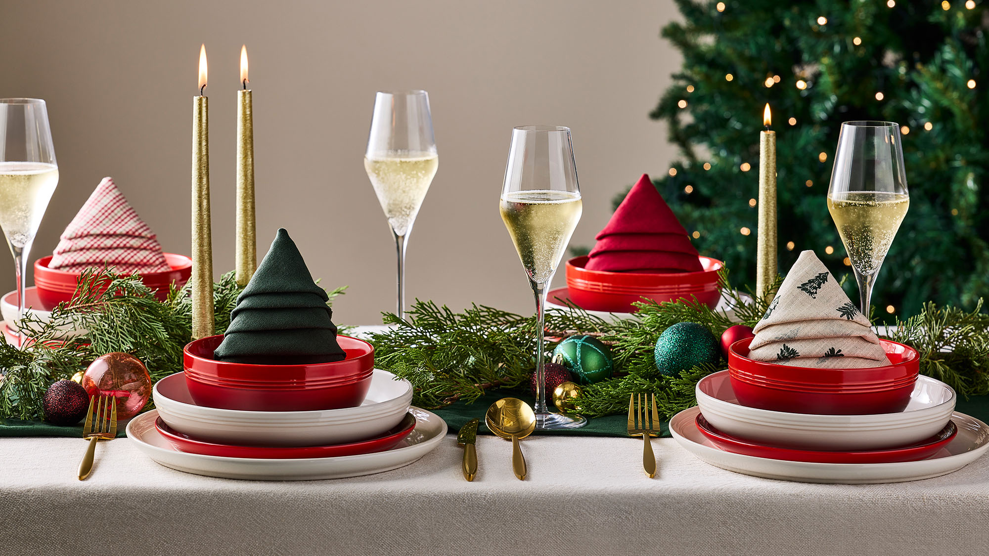 Festive dining table with red and white plates, folded napkins, candles, glasses of champagne, and Christmas decorations, set near a Christmas tree.