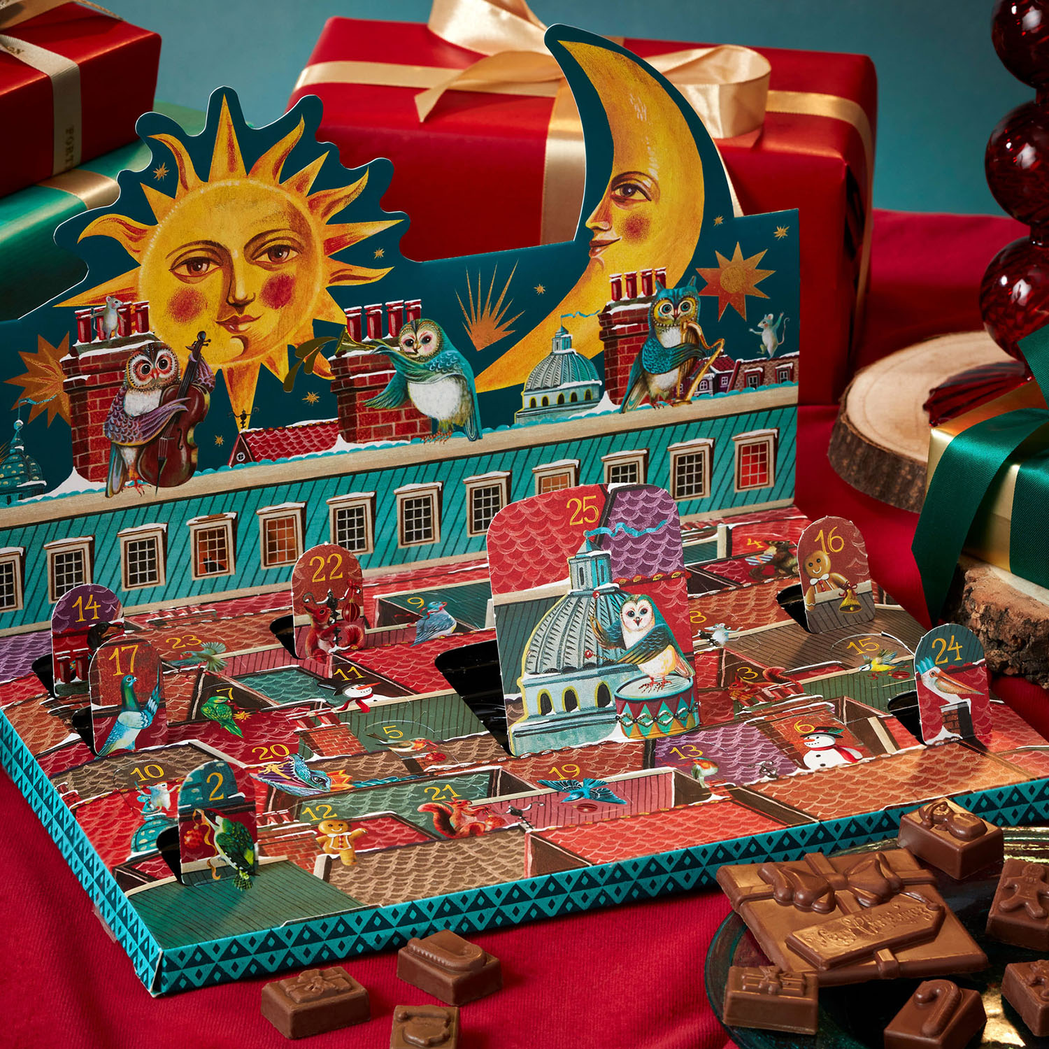 A festive advent calendar with open doors, revealing chocolates, surrounded by wrapped gifts on a red cloth.