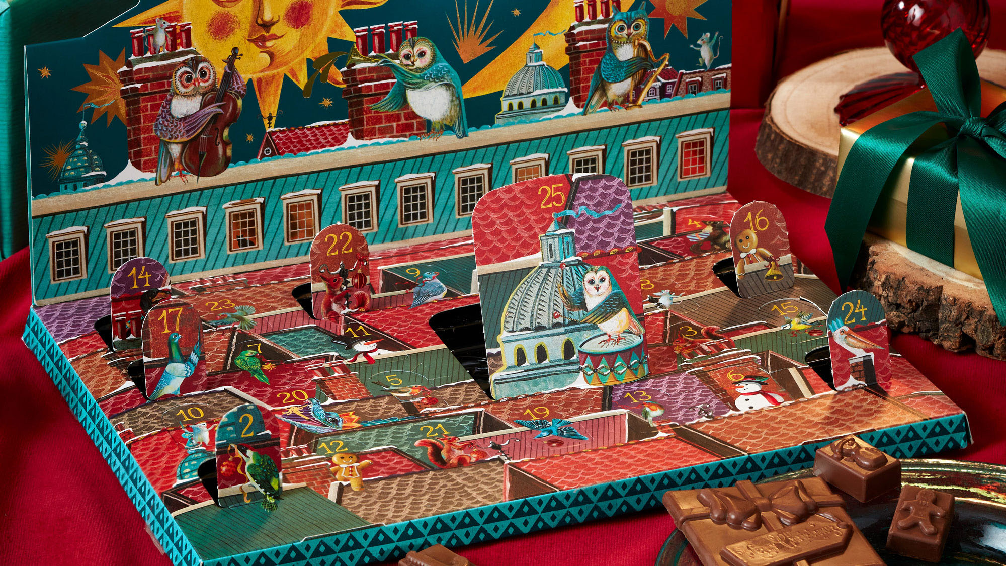 A festive advent calendar with open doors, revealing chocolates, surrounded by wrapped gifts on a red cloth.