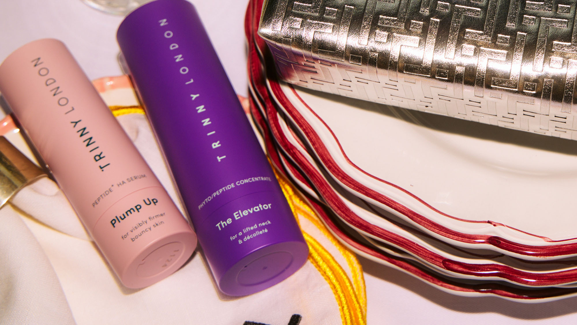 A table with skincare products, a metallic clutch, stacked plates, and a glass of water.