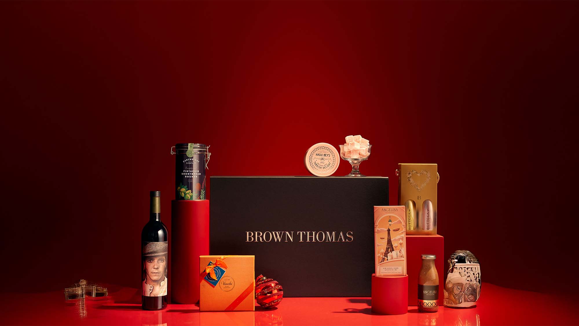A selection of luxury gift items are arranged against a red background, featuring a prominent Brown Thomas box and various gourmet products.