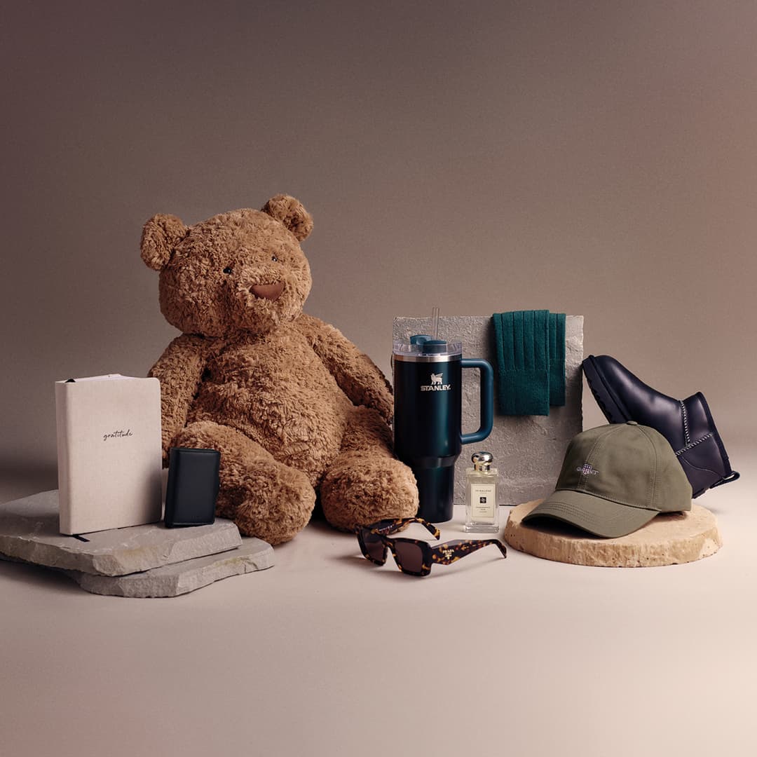 A teddy bear sits among various items: notebooks, tumblers, sunglasses, cologne, a cap, scarf, and a pair of black shoes.
