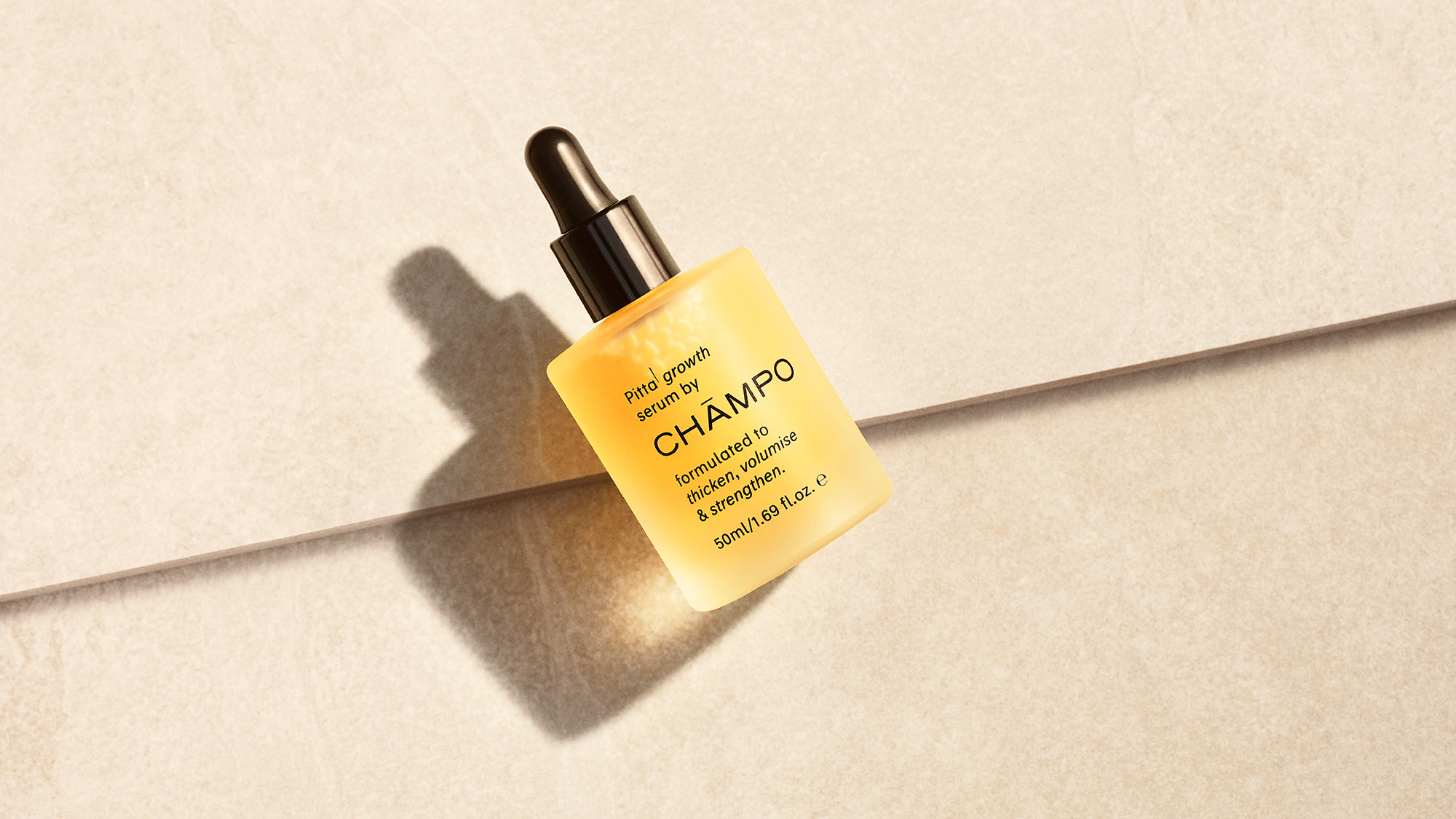 A bottle of Champo hair oil with a dropper lies on a light, textured surface, casting a shadow.