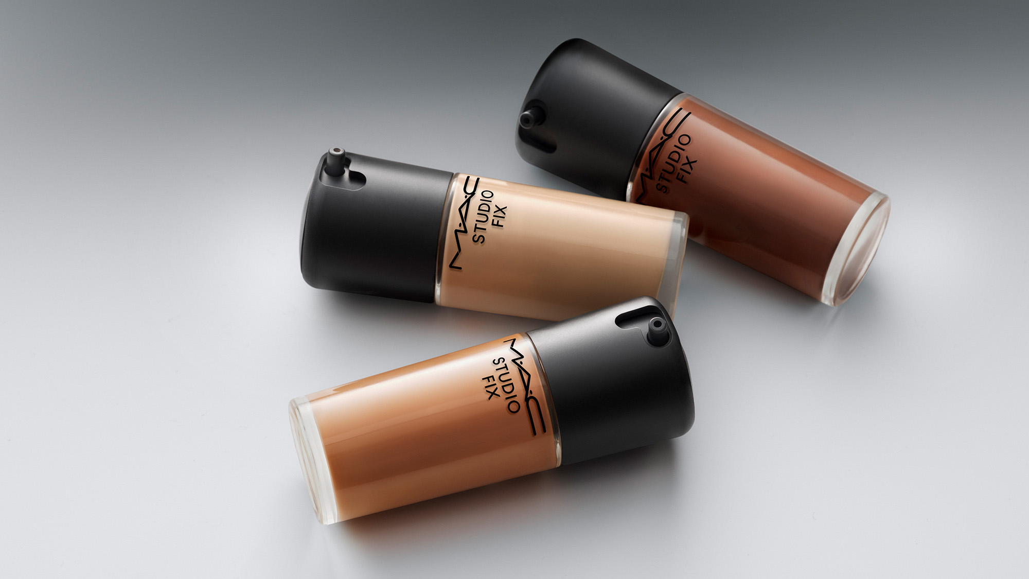 Three MAC Studio Fix liquid foundation bottles in different shades on a smooth surface.