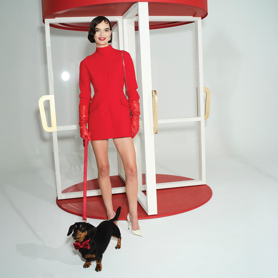 A woman in a red dress and gloves stands in a revolving door with a small black and brown dog on a red leash.