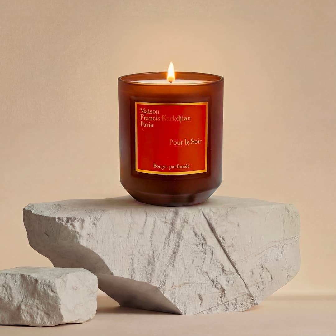 A lit, brown glass candle with a red label sits on textured stone slabs against a neutral background.