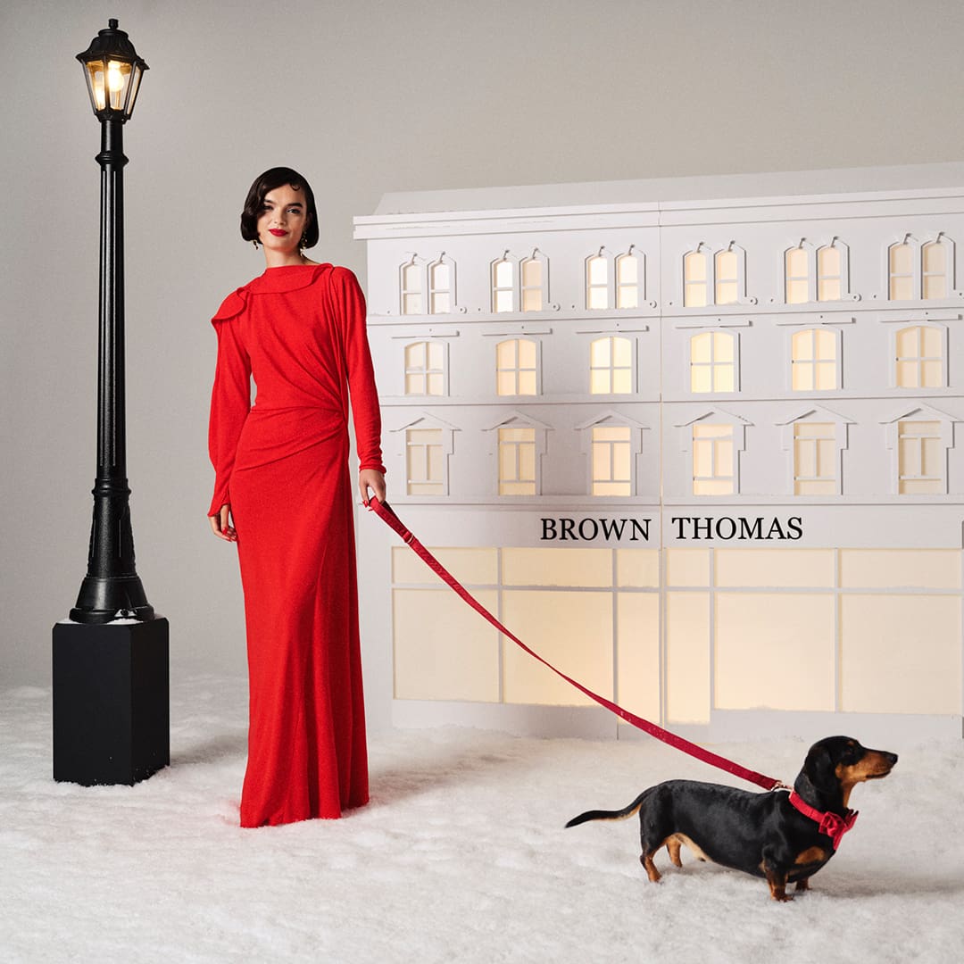 A person in a red dress stands with a dachshund on a leash. They are in front of a model building labeled 'Brown Thomas,' flanked by two lampposts.
