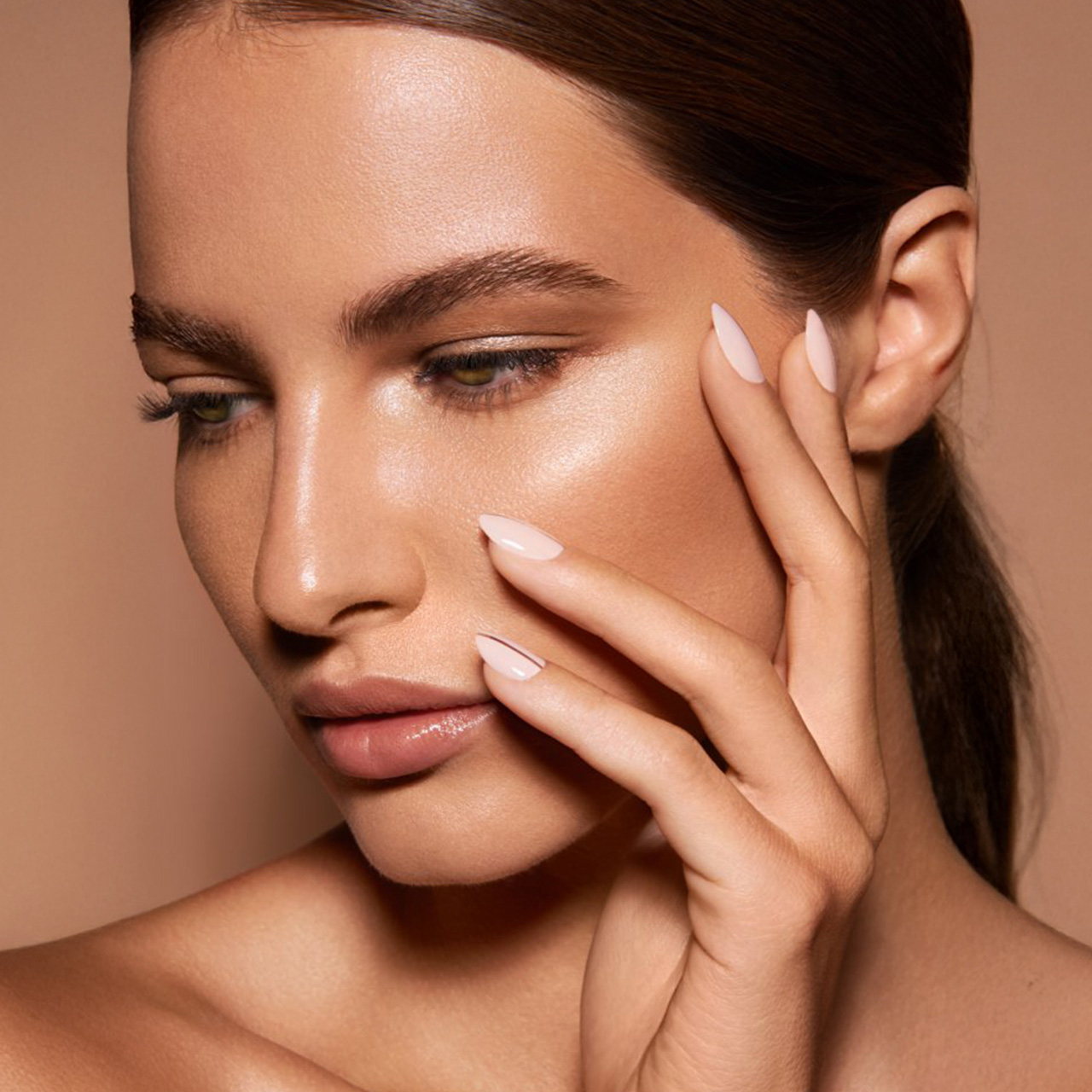 A woman with smooth skin and light pink nails poses with her hand near her face against a neutral background.