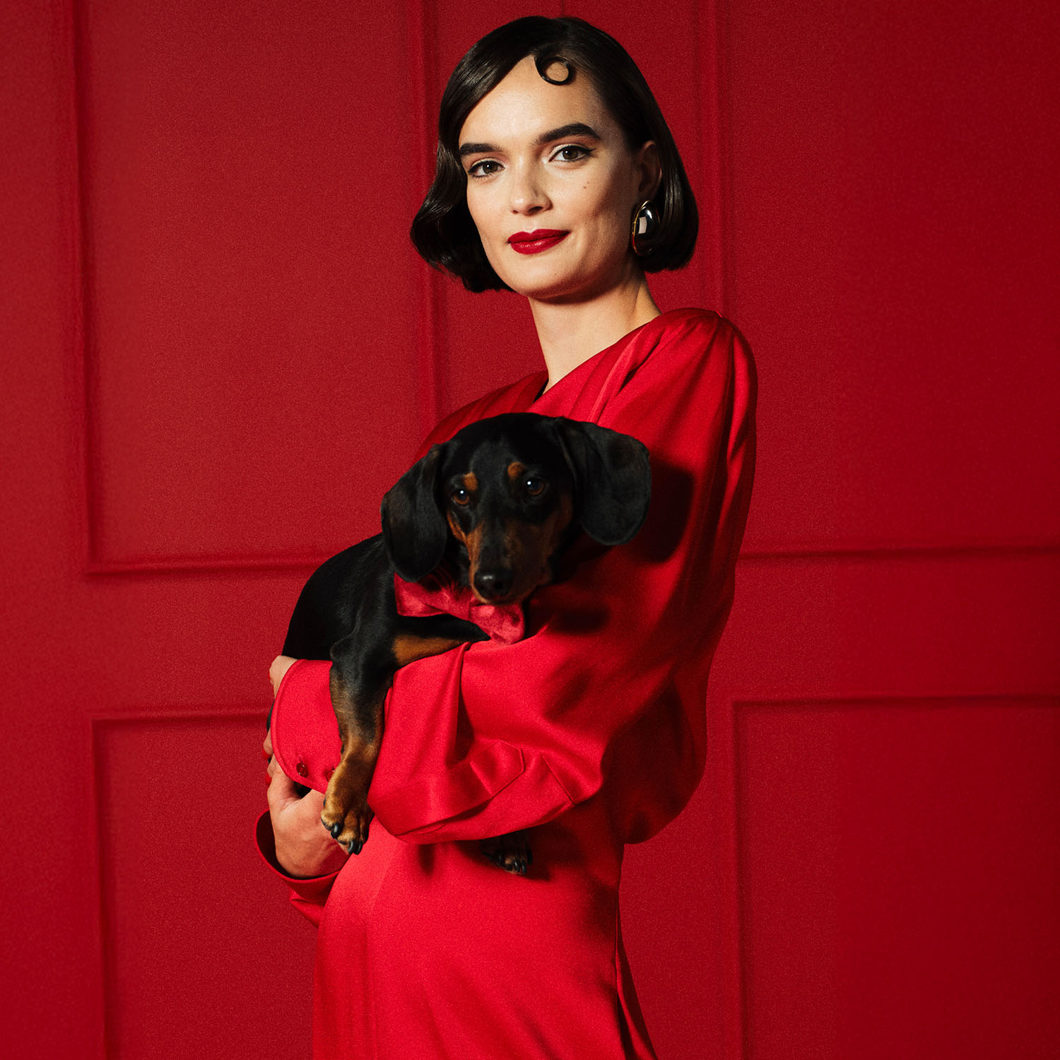 A woman in a red outfit holding a small black and tan dog against a red background.