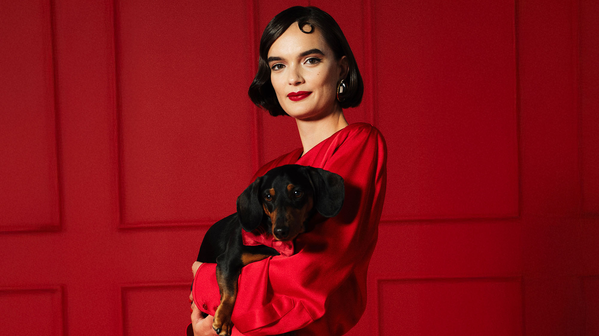 A woman in a red outfit holding a small black and tan dog against a red background.