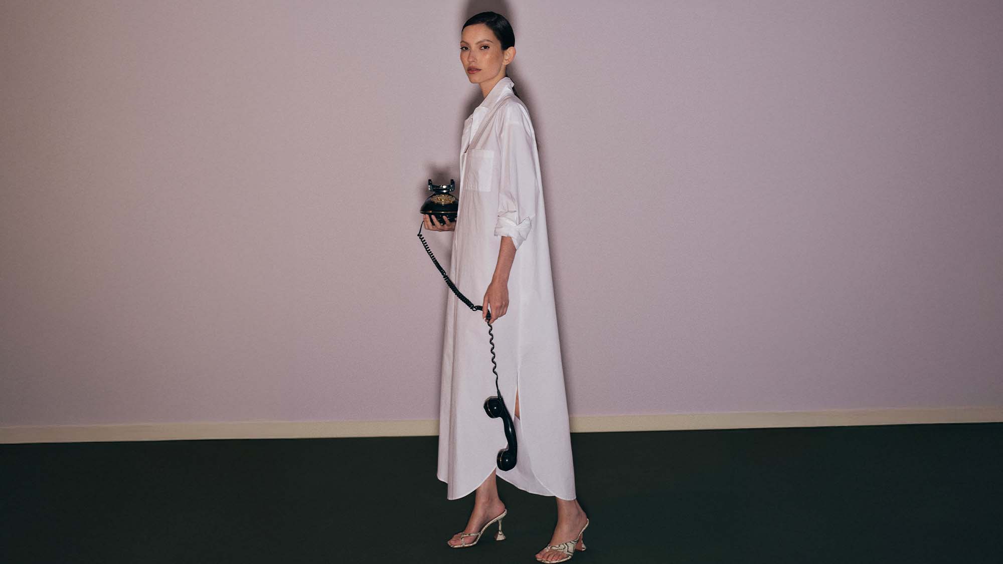 A woman in a long white shirt dress holds a vintage telephone, standing on a dark floor against a light purple wall.