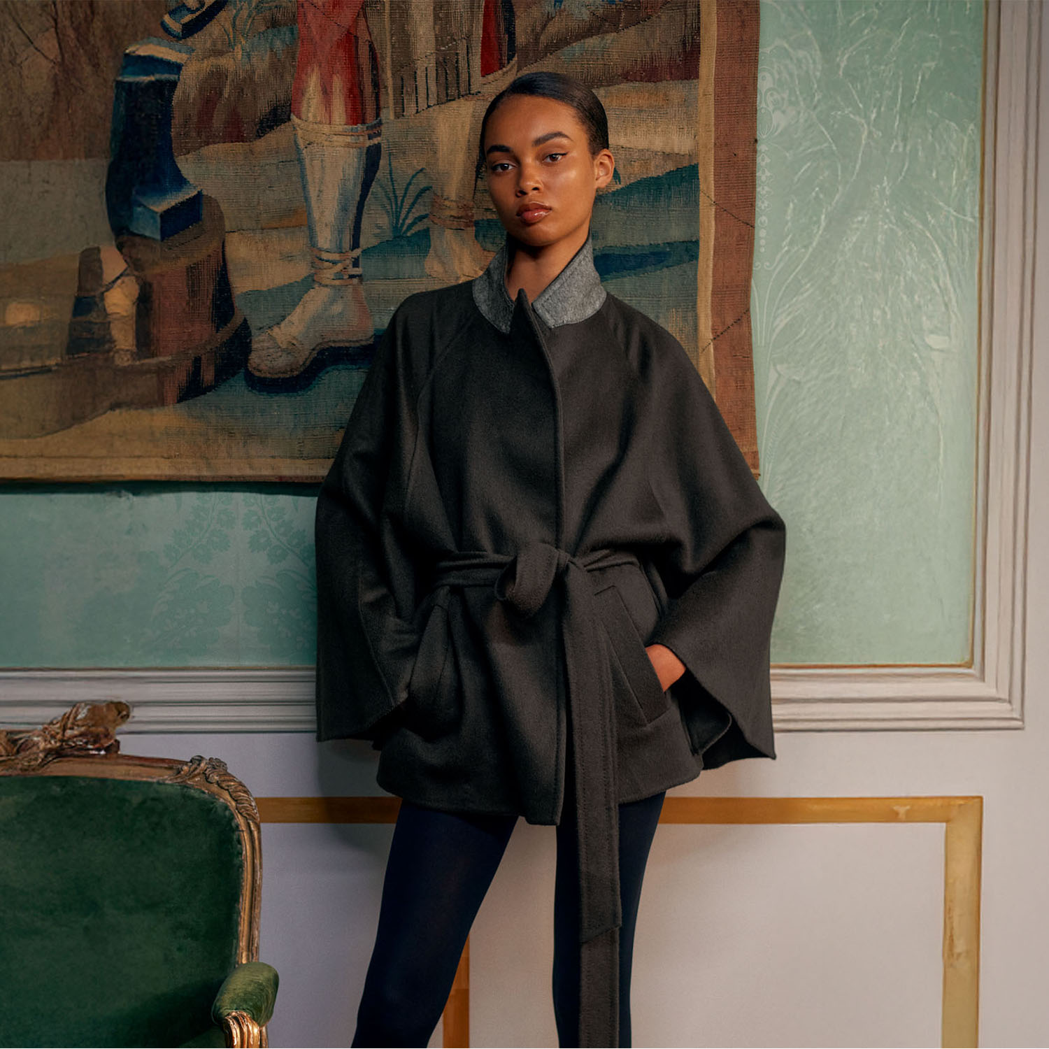 A person in a dark, belted coat stands against a tapestry wall, with a green chair nearby.
