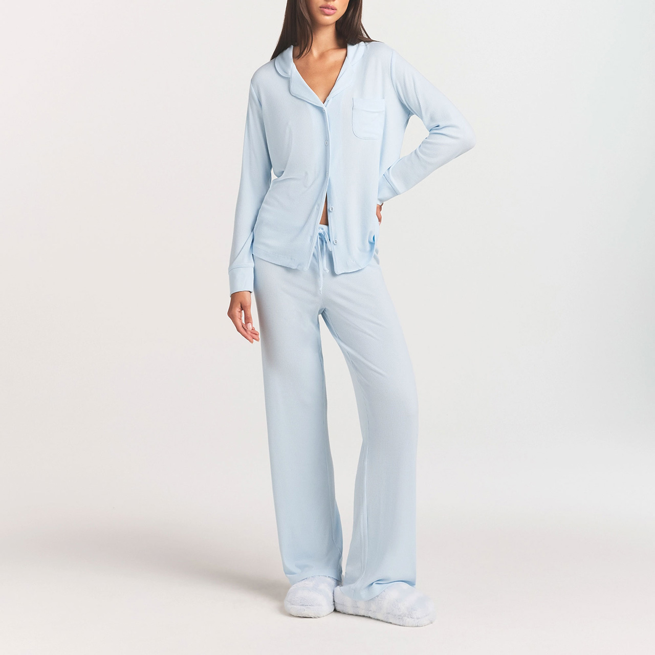 A person wearing light blue pajamas and white slippers stands with one hand in a pocket against a plain background.